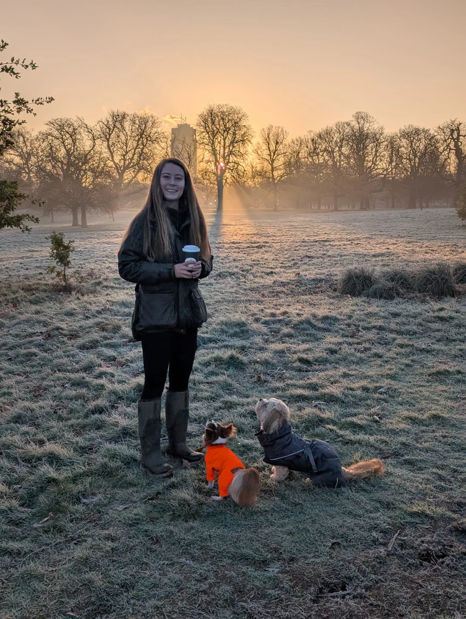 A smiling woman holding a coffee cup with two little dogs sitting looking at her.