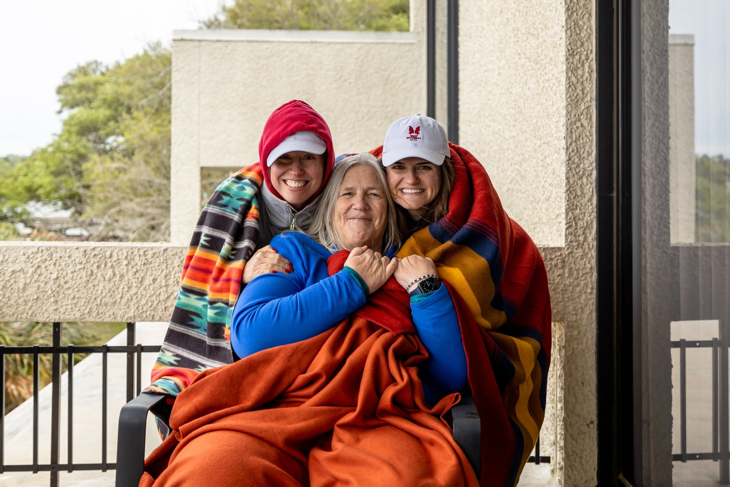 Three women smiling and hugging outdoors on a balcony, wrapped in colorful blankets, with one woman seated and two standing behind her, all wearing hats and casual clothing.