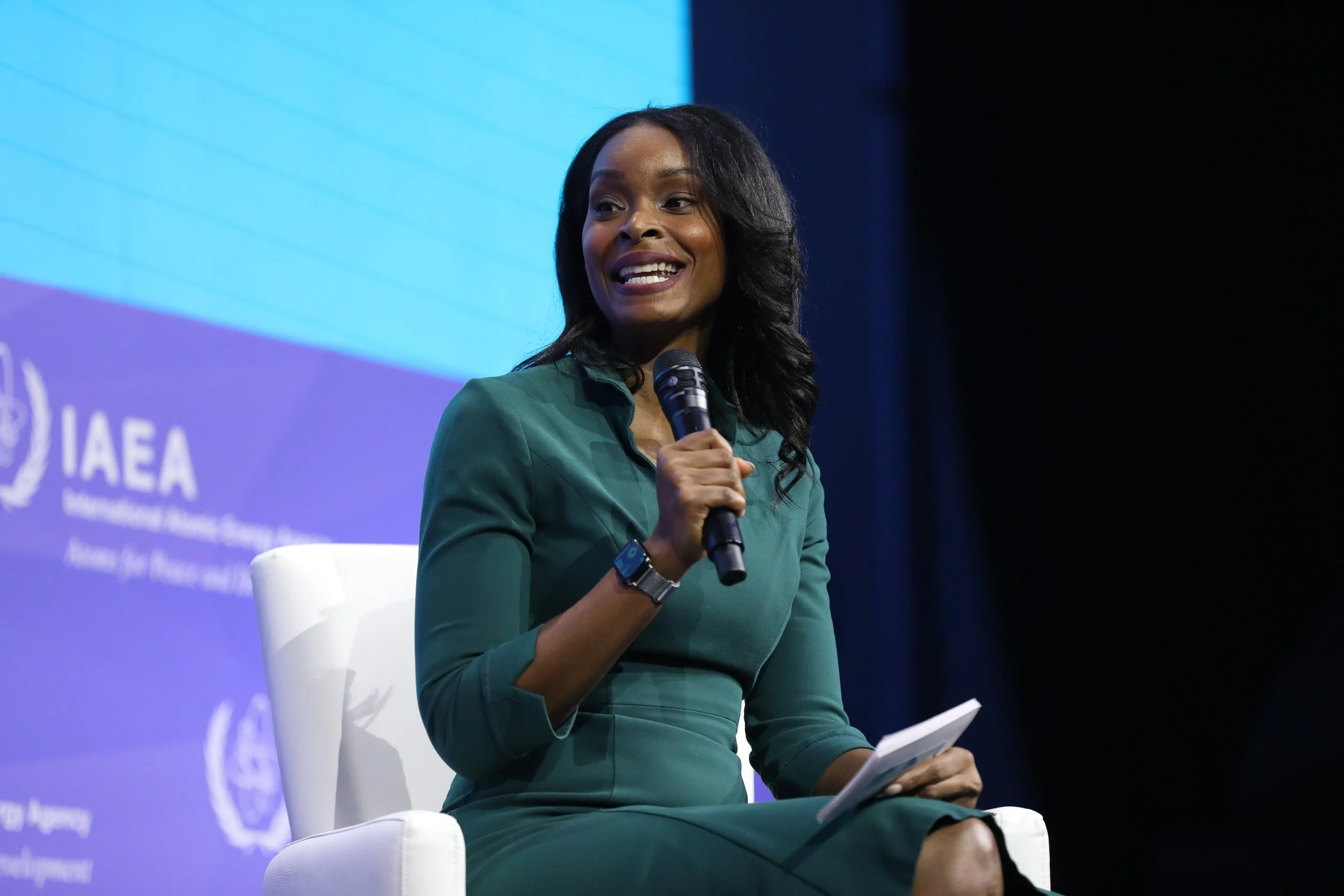 African American woman in green dress speaking at a conference, holding a microphone and notes, seated on a white chair with a blue background displaying the IAEA logo.