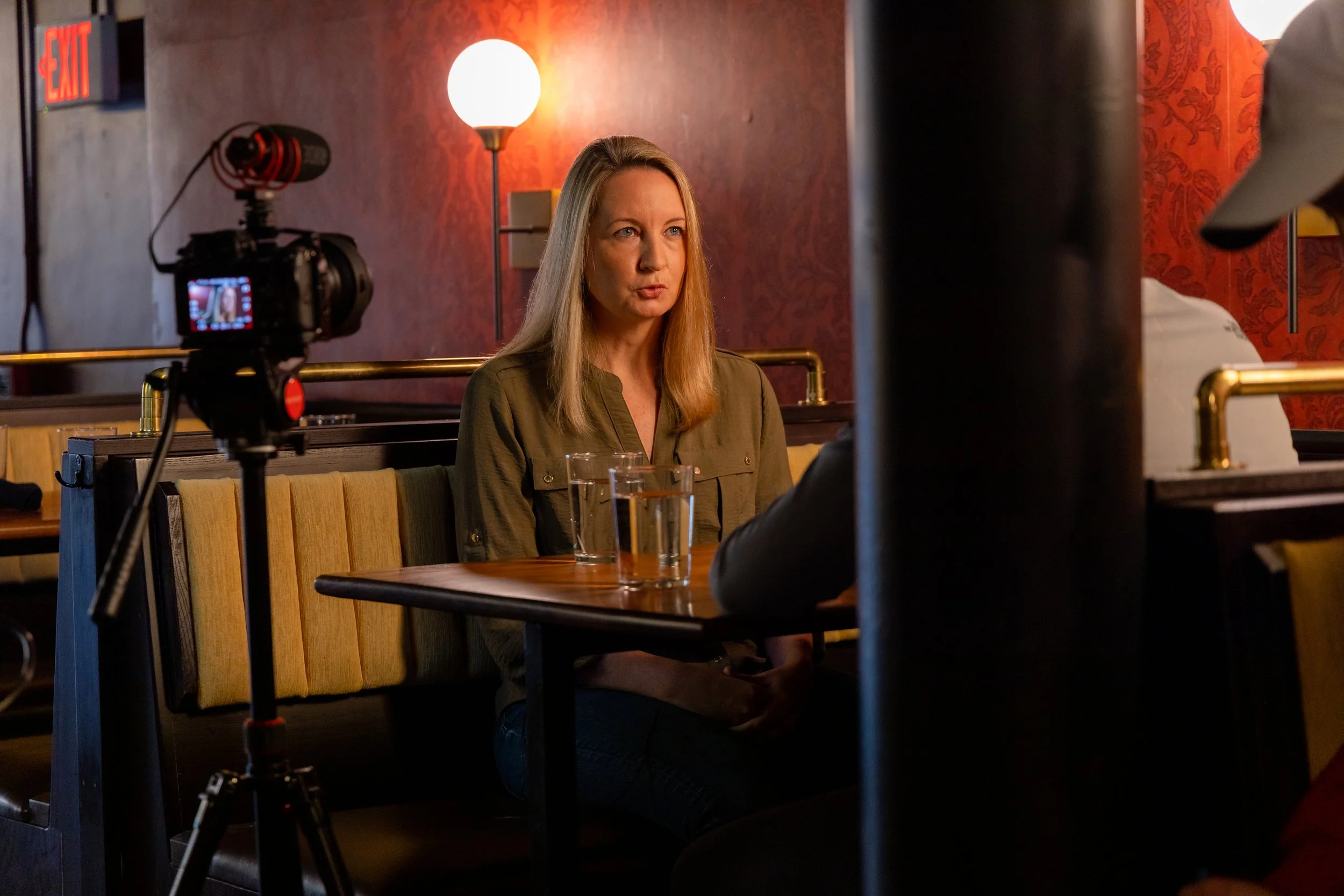 A woman with long blonde hair sitting at a booth in a restaurant or bar, being recorded on a camera while having a conversation with a person to her right. The scene is lit with warm lighting, with a red patterned wall and a lamp in the background.