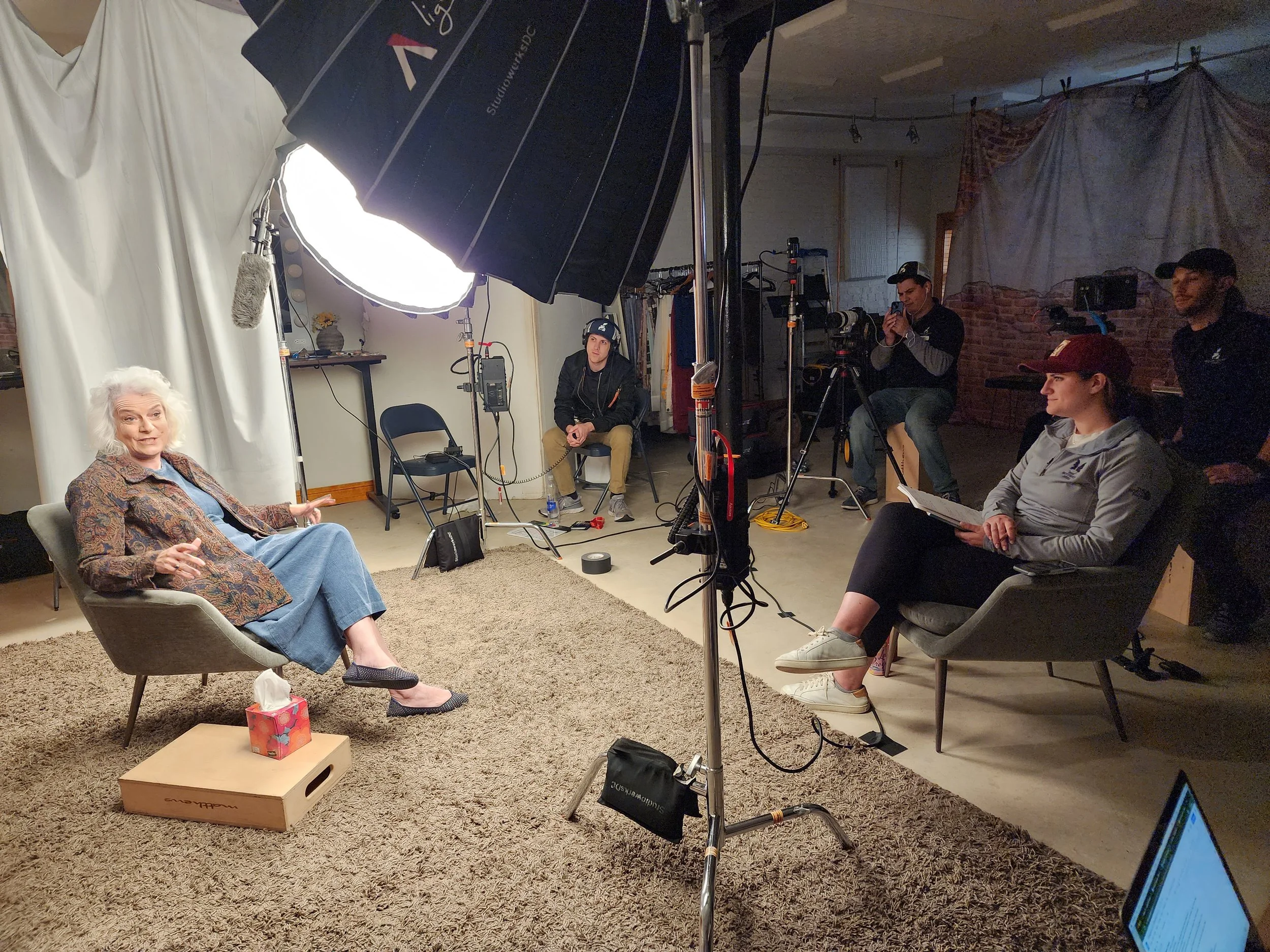 A woman in a brown patterned blazer and blue dress sitting in a modern armchair during a filming or interview session inside a studio, with professional lighting and crew members working around her.