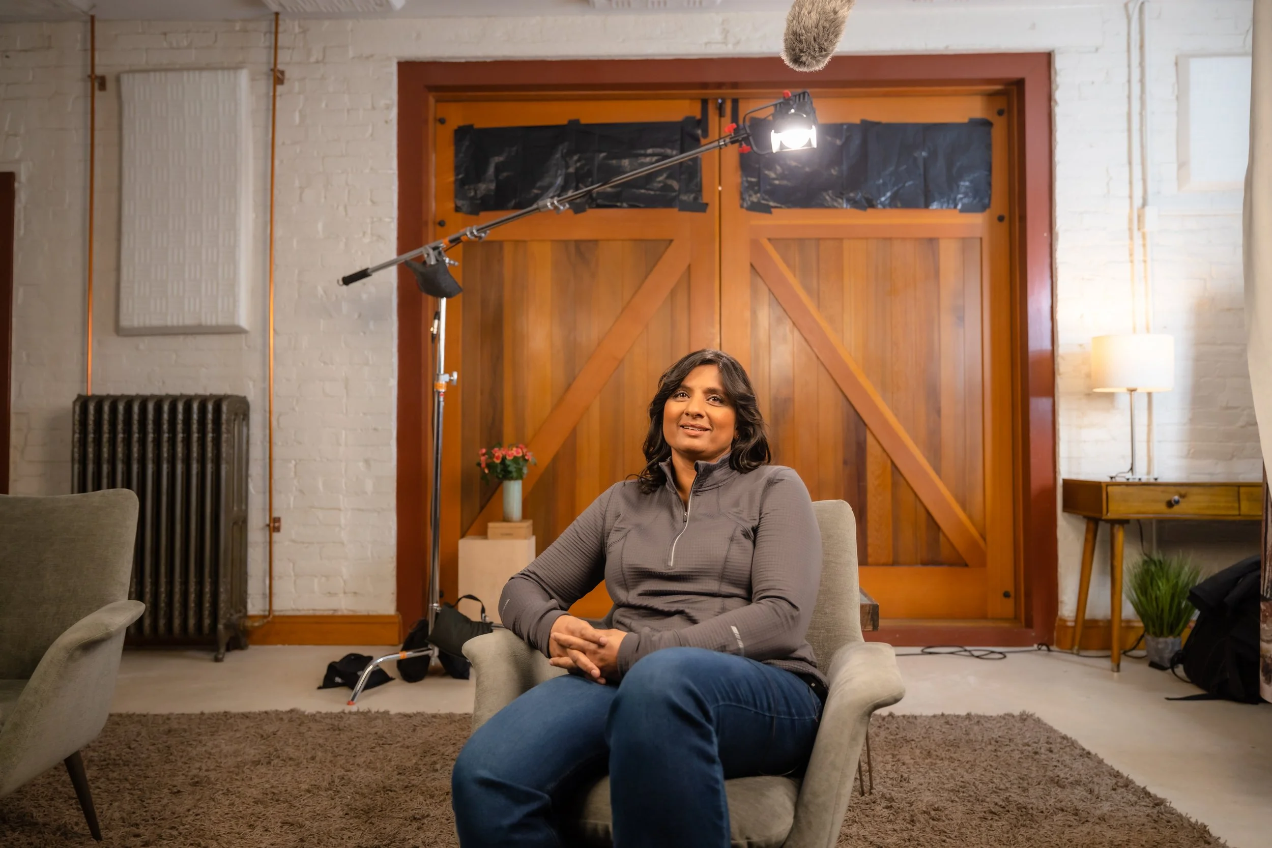 A woman sitting on a beige armchair in a room with a wooden barn-style door, a lamp, a small table with a potted plant, and lighting equipment overhead, during a video or photo shoot.