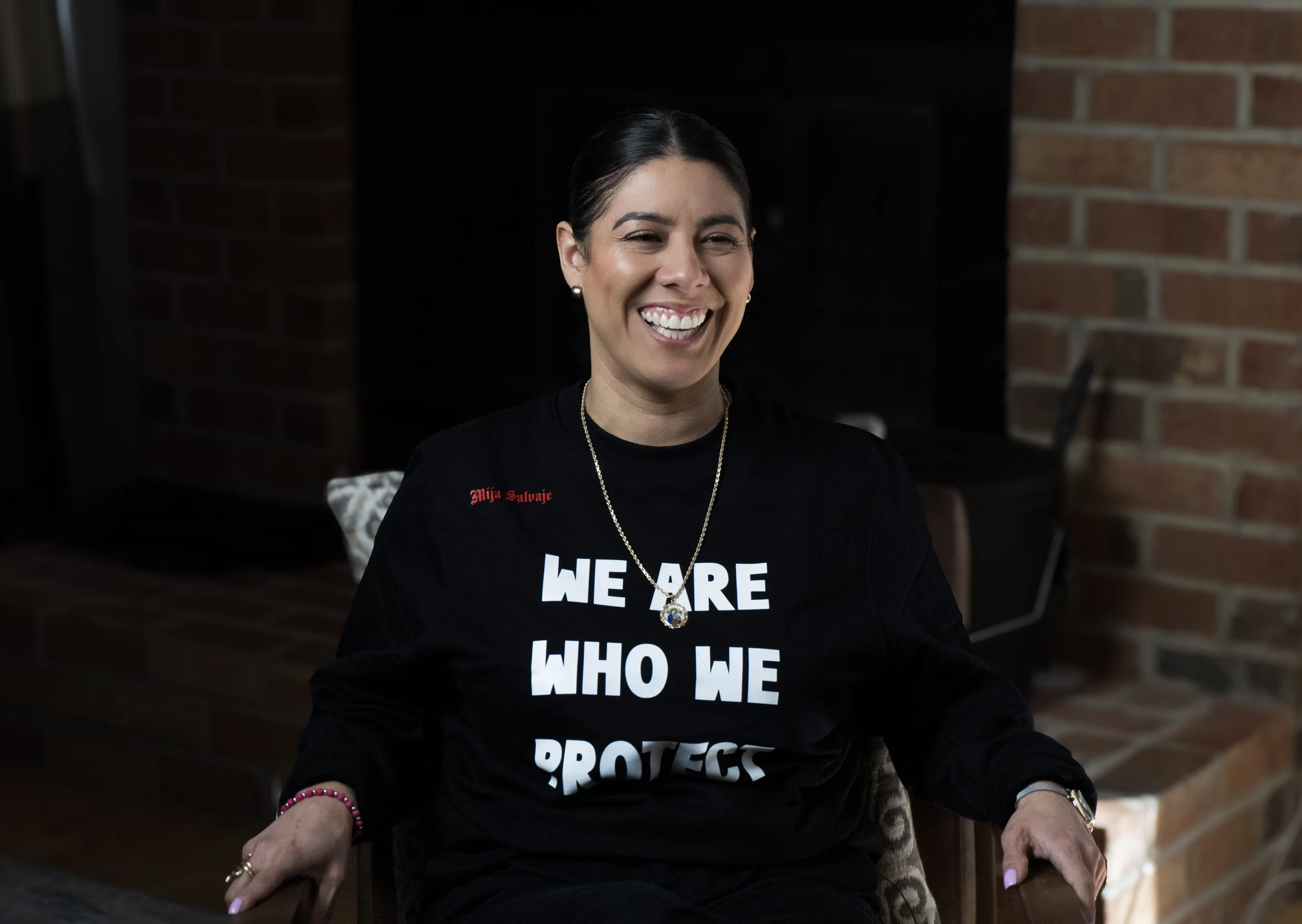 A woman sitting in a chair, smiling, wearing a black shirt with white text that reads "We are who we protect," and a necklace with a pendant, in front of a brick fireplace.