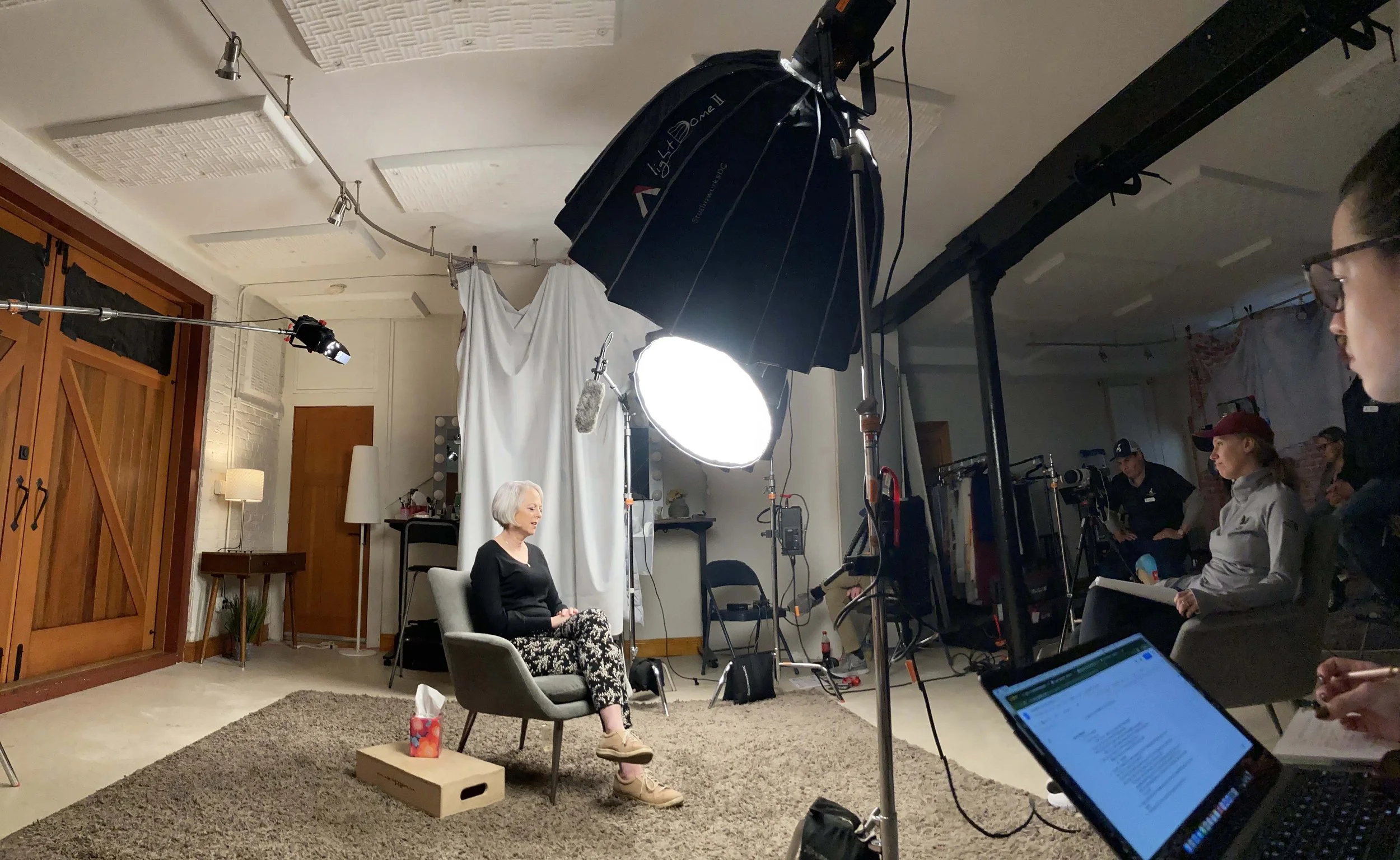 A woman with gray hair sitting on a light gray chair in a recording studio or interview set, with a large professional light overhead. Behind her is a white curtain, and various equipment and crew members are visible to the right.