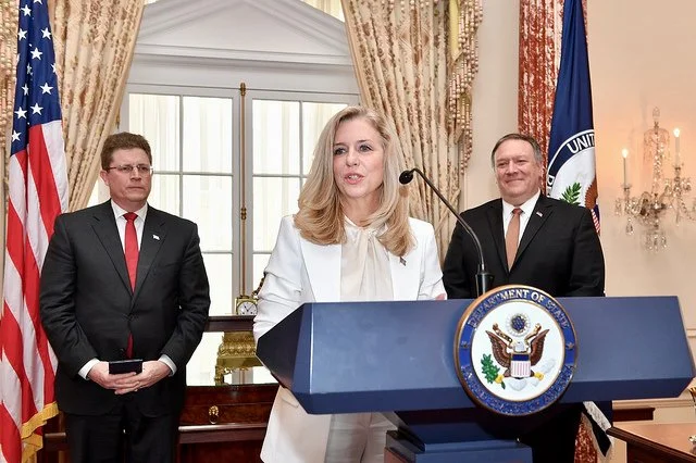 A woman is speaking at a podium with the Seal of the U.S. Department of Justice, flanked by two men in suits. The setting is a formal room with an American flag and a Department of Justice flag in the background.