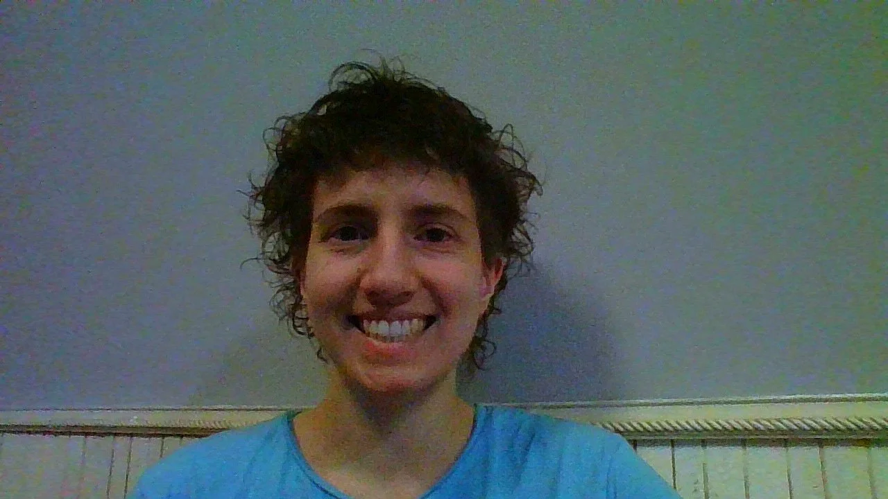 A person with short, curly hair smiling in front of a plain wall.