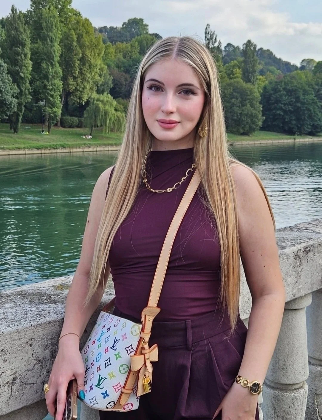Young woman with long blonde hair standing outdoors near water, wearing a sleeveless dark purple top, gold jewelry, and carrying a colorful designer bag, with trees and a cloudy sky in the background.