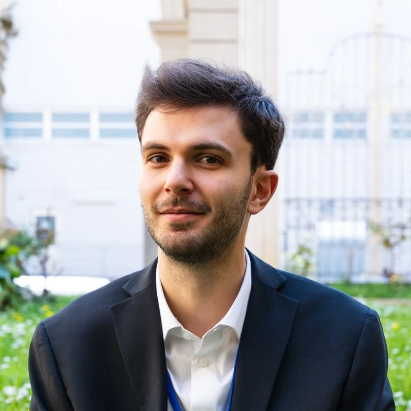 Headshot of a young man with dark hair and a beard, wearing a black suit and white shirt, standing outdoors in a residential area.