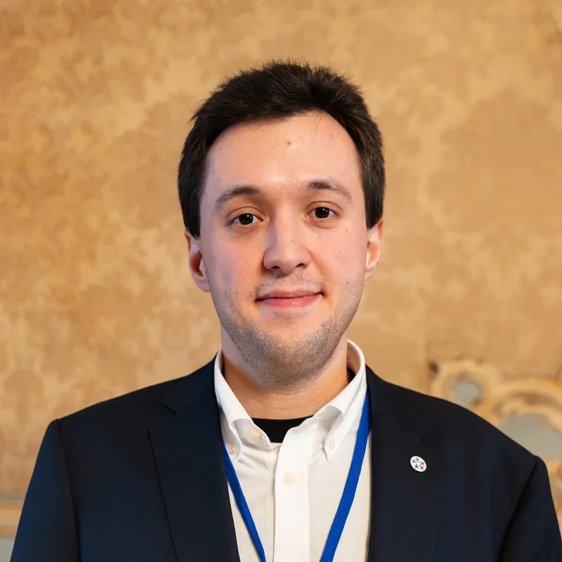 Portrait of a young man in a dark suit with white shirt, wearing a blue lanyard, standing in front of a beige textured wall.