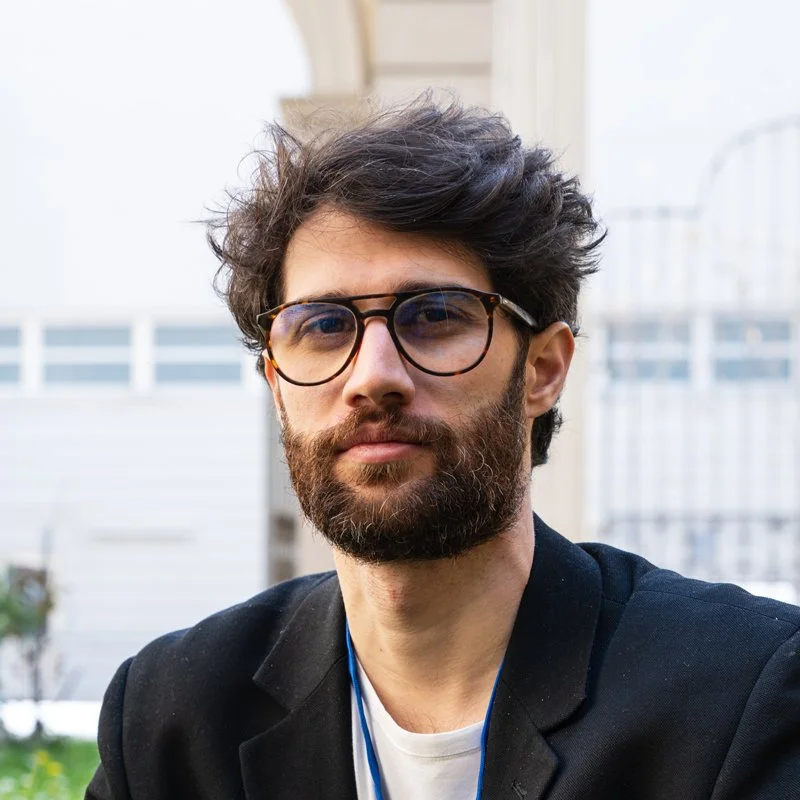 A man with dark, curly hair and a beard wearing glasses, a black blazer, and a white shirt outdoors in front of a house.