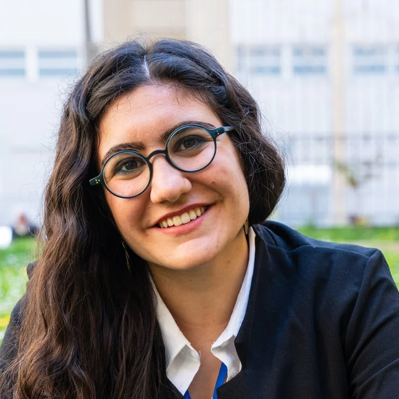 Smiling woman with long wavy hair, wearing round glasses and a black blazer, outdoors with a city background.