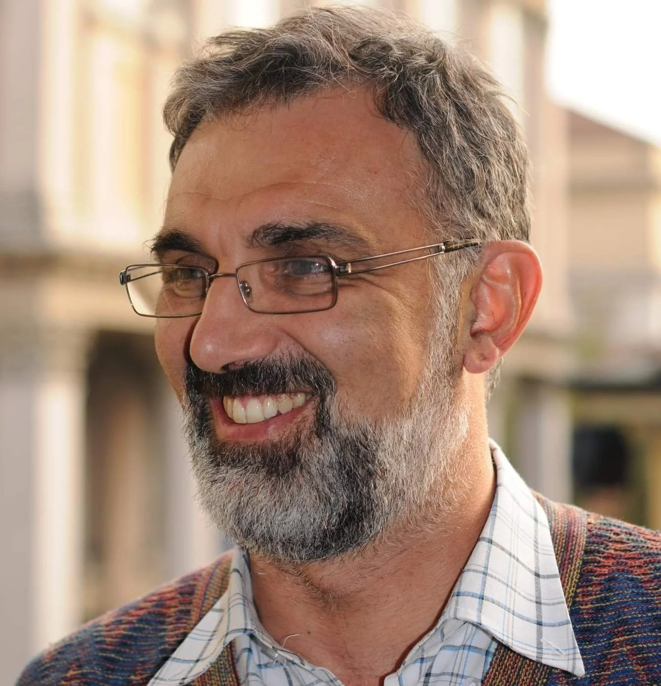 A smiling middle-aged man with glasses, a beard, and wearing a plaid shirt and sweater.