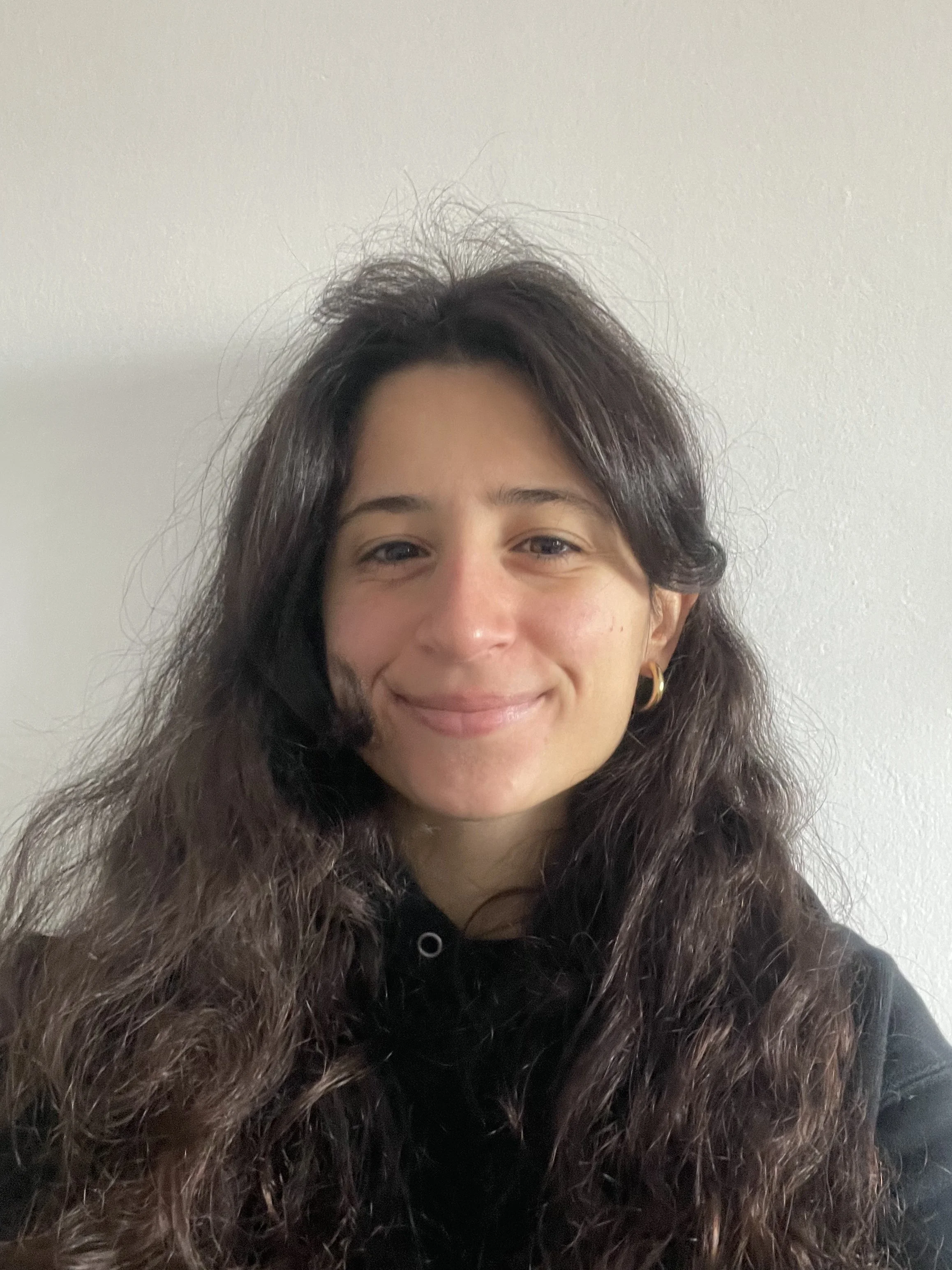 A woman with long, dark wavy hair and hoop earrings, smiling at the camera in front of a plain white wall.