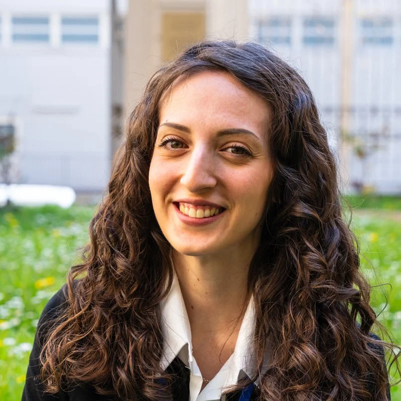 Portrait of a woman with long, curly brown hair, smiling outdoors with greenery and a building in the background.