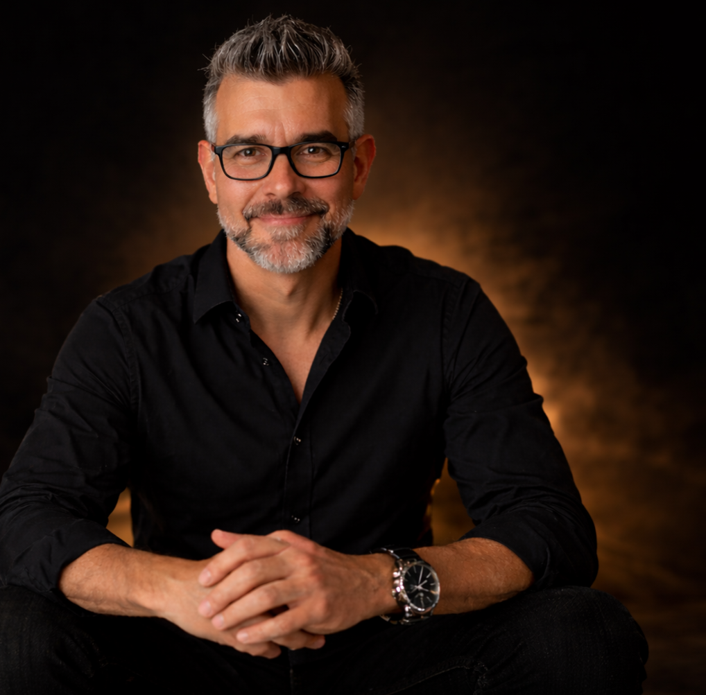 Ein Mann mit Brille, grauem Haar und Bart, trägt ein schwarzes Hemd, sitzt vor einem dunklen Hintergrund mit warmem Licht. Er lächelt leicht und hat die Hände zusammengefaltet.