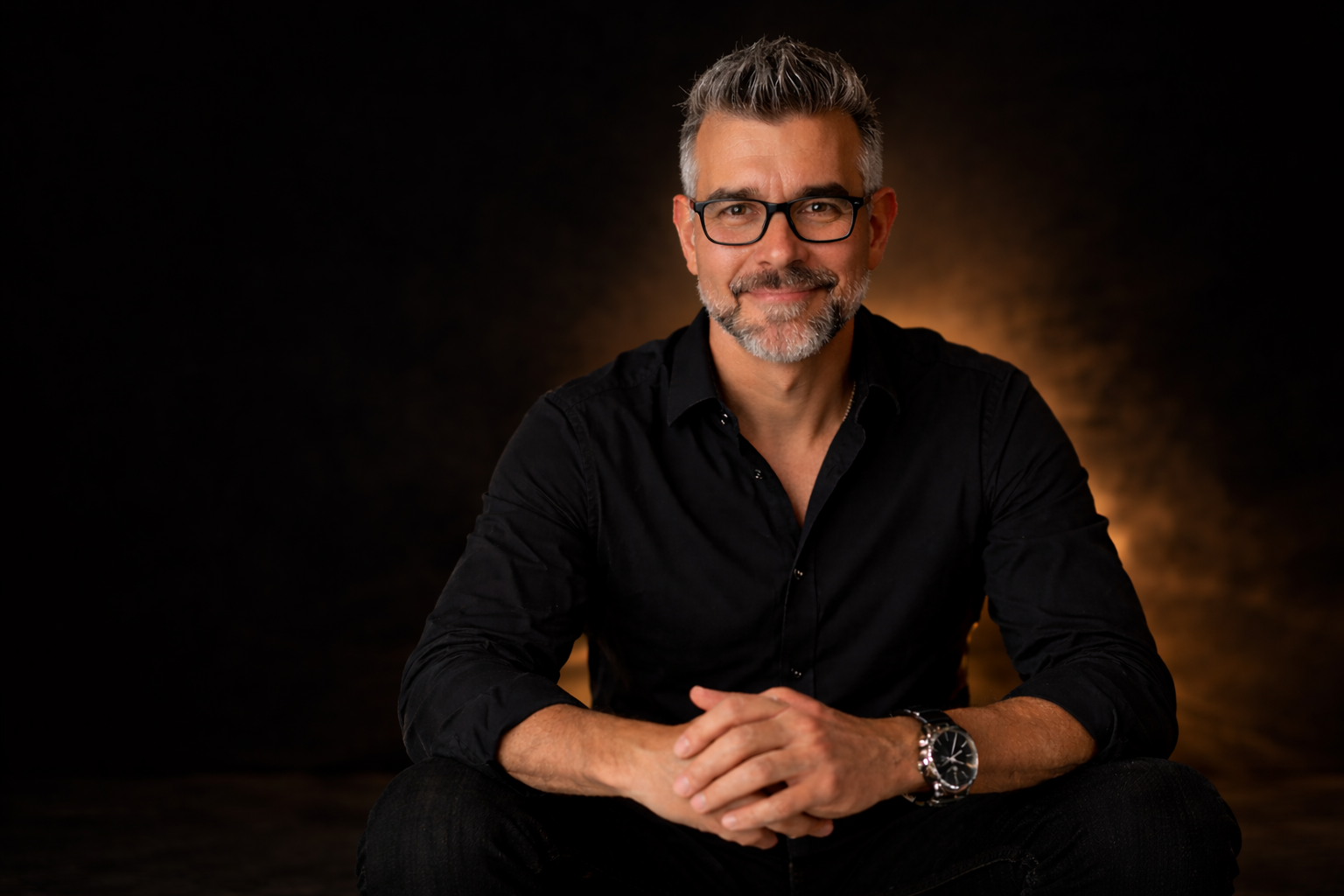 Porträt eines lächelnden Mannes mit Brille, grauem Haar und Bart, in schwarzem Hemd, sitzend mit verschränkten Händen vor dunklem Hintergrund