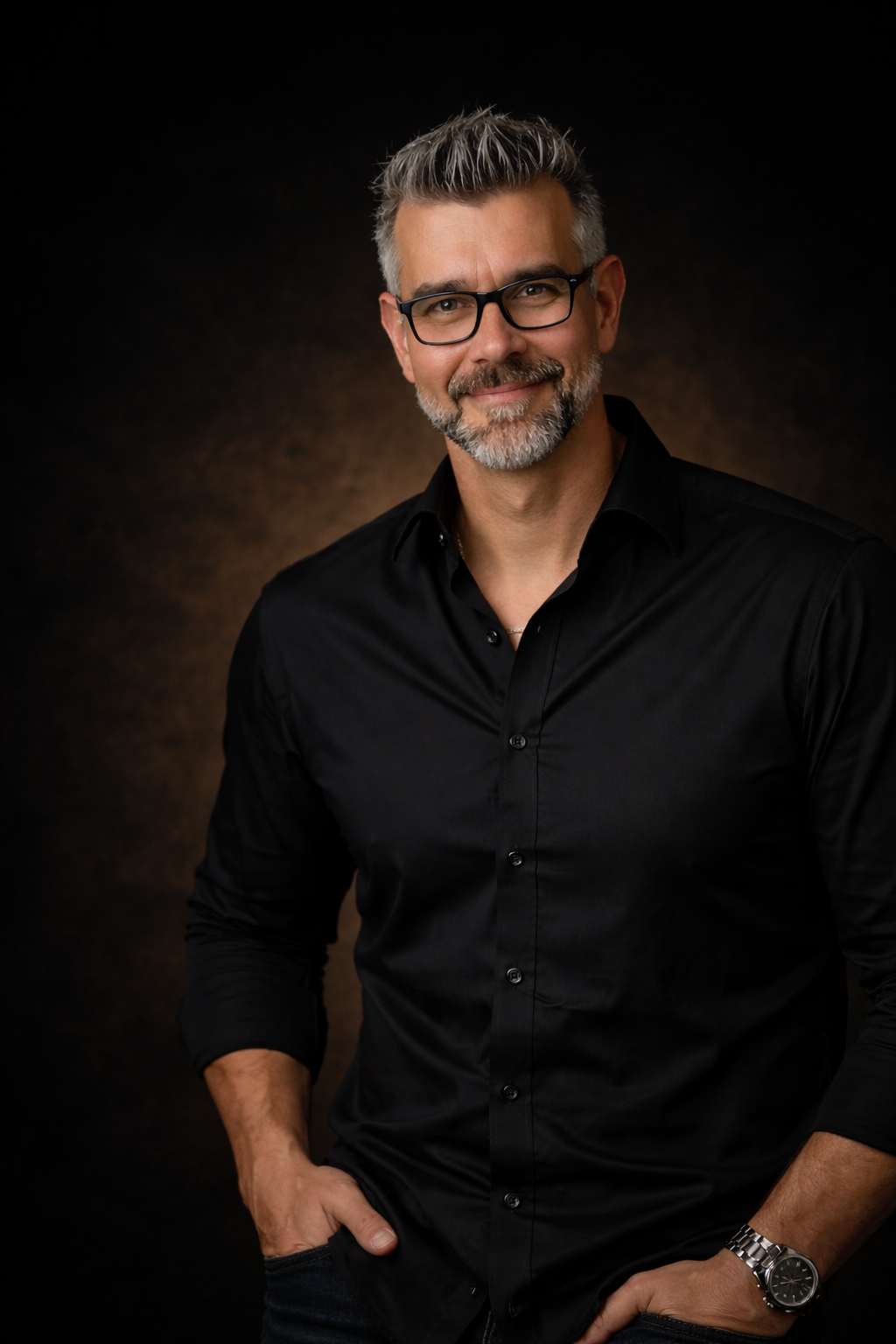 Ein Mann mit grauem Haar, Brille und Bart, trägt ein schwarzes Hemd, lächelt und hat die Hände in den Taschen, vor dunklem Hintergrund.