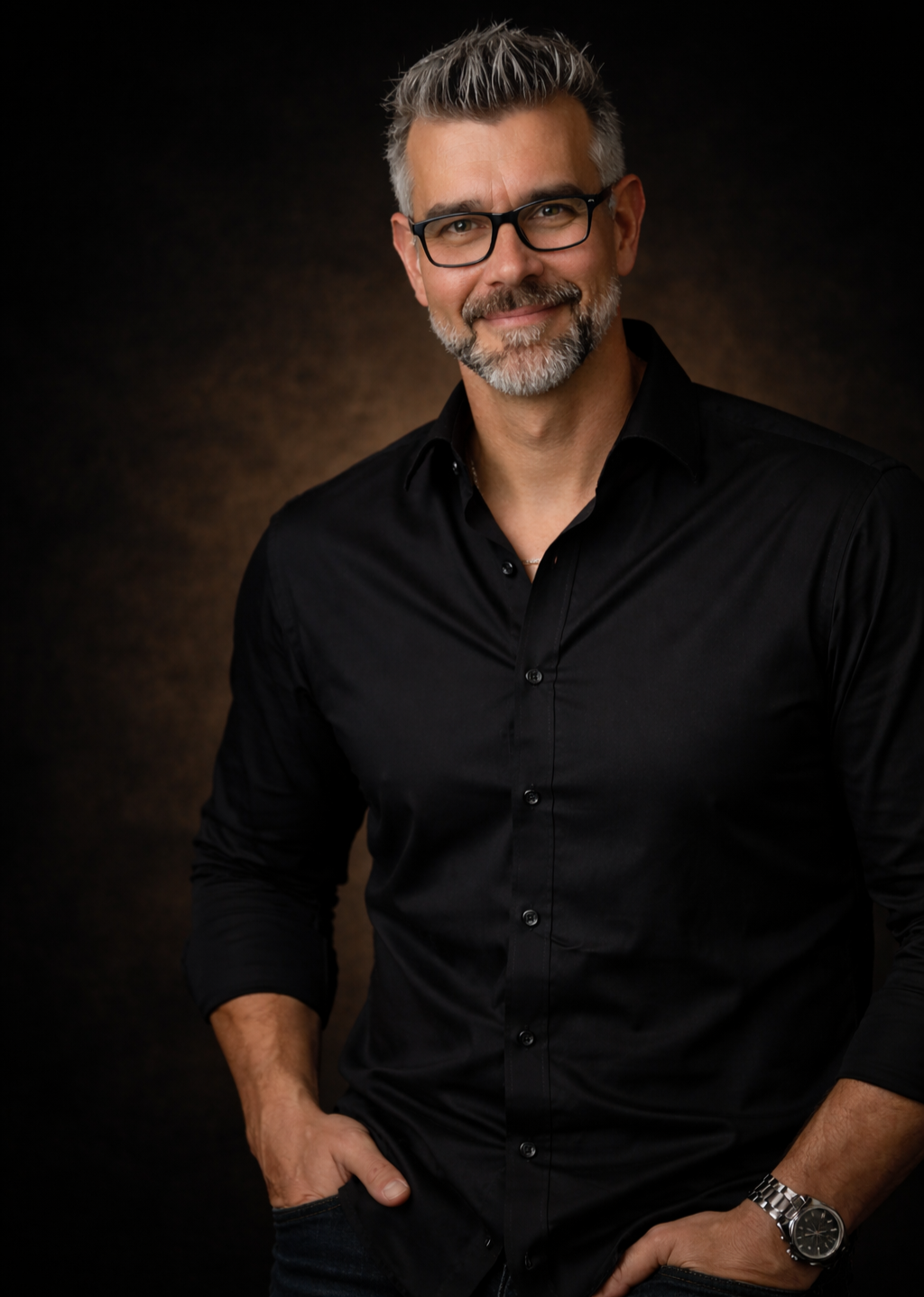 Ein Mann mit Brille, grauem Haar und Bart trägt ein schwarzes Hemd und posiert lächelnd vor dunklem Hintergrund.