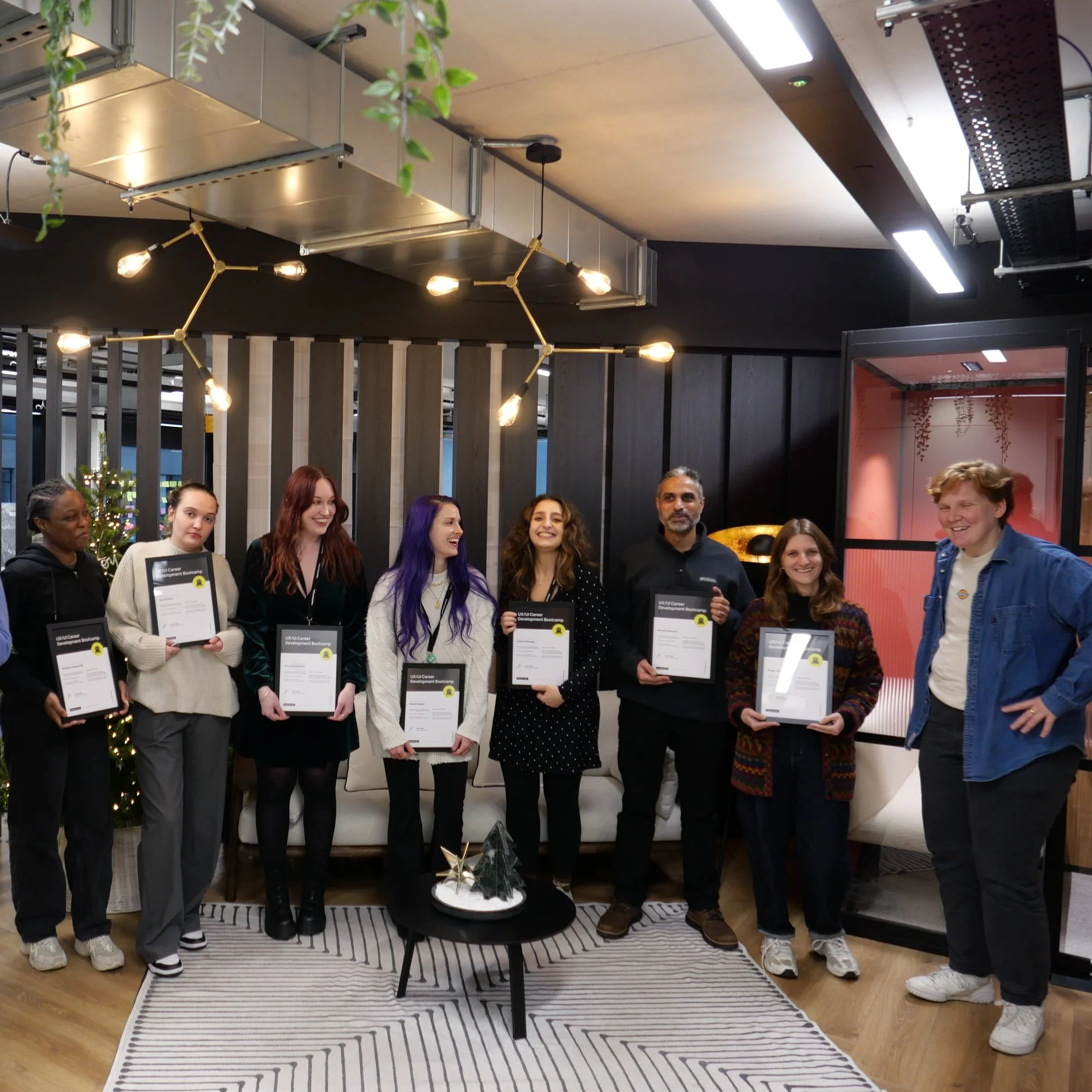 Group of eight individuals standing in a modern indoor space holding certificates, celebrating an achievement. They are smiling and dressed casually, with a small decorated Christmas tree and festive decorations in the background.