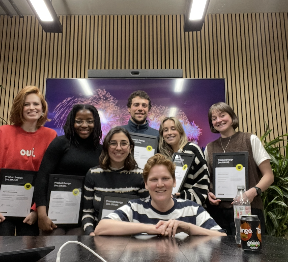 Group of seven young adults in an office celebrating, some holding awards, in front of a large screen with fireworks display, on a wooden wall, with a potted plant and drinks on the table.