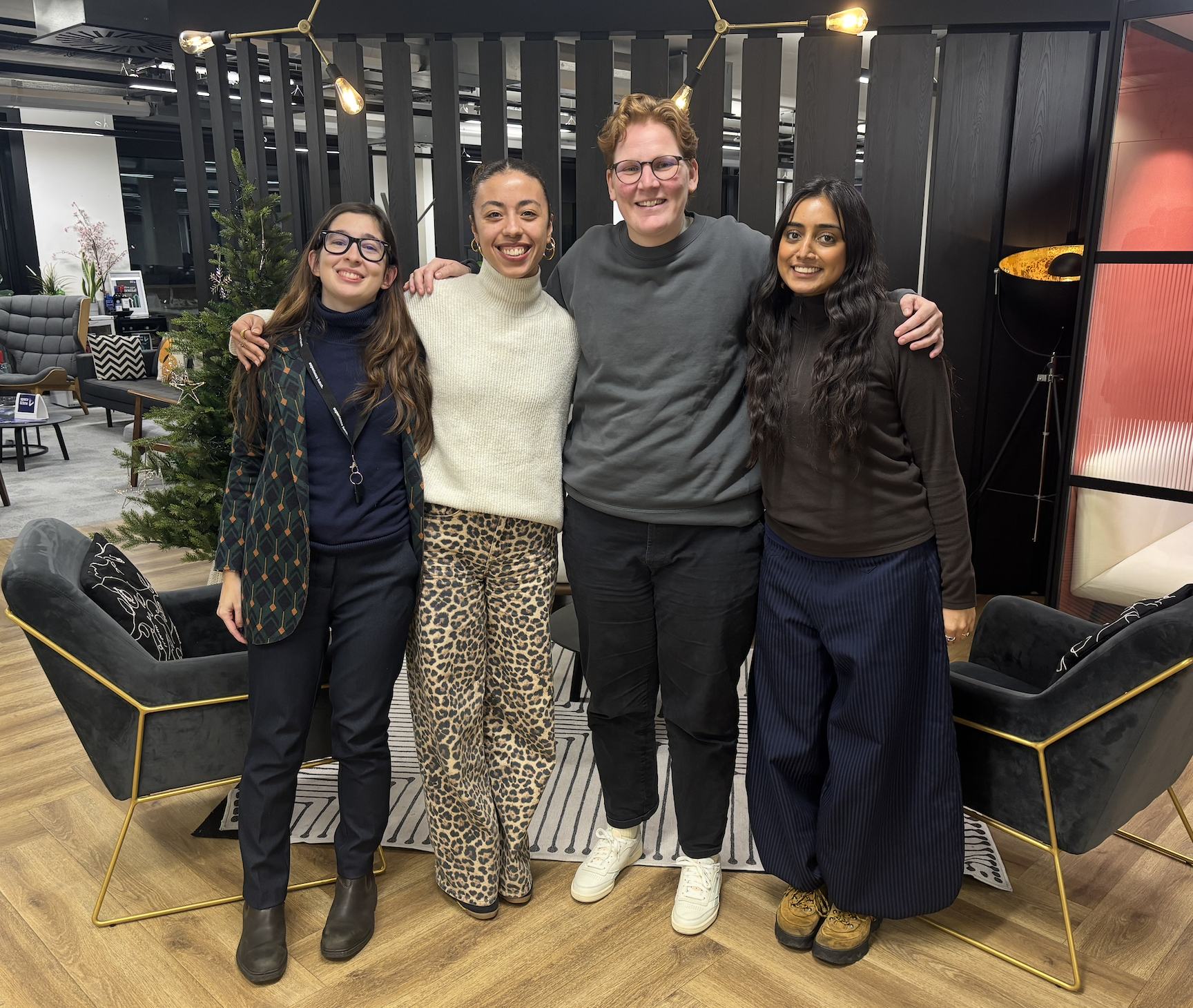Four people standing closely together in an office lobby, smiling at the camera. The group consists of two women and one man, with another woman on the far right. The environment features modern furniture, a Christmas tree, and decorative lighting.