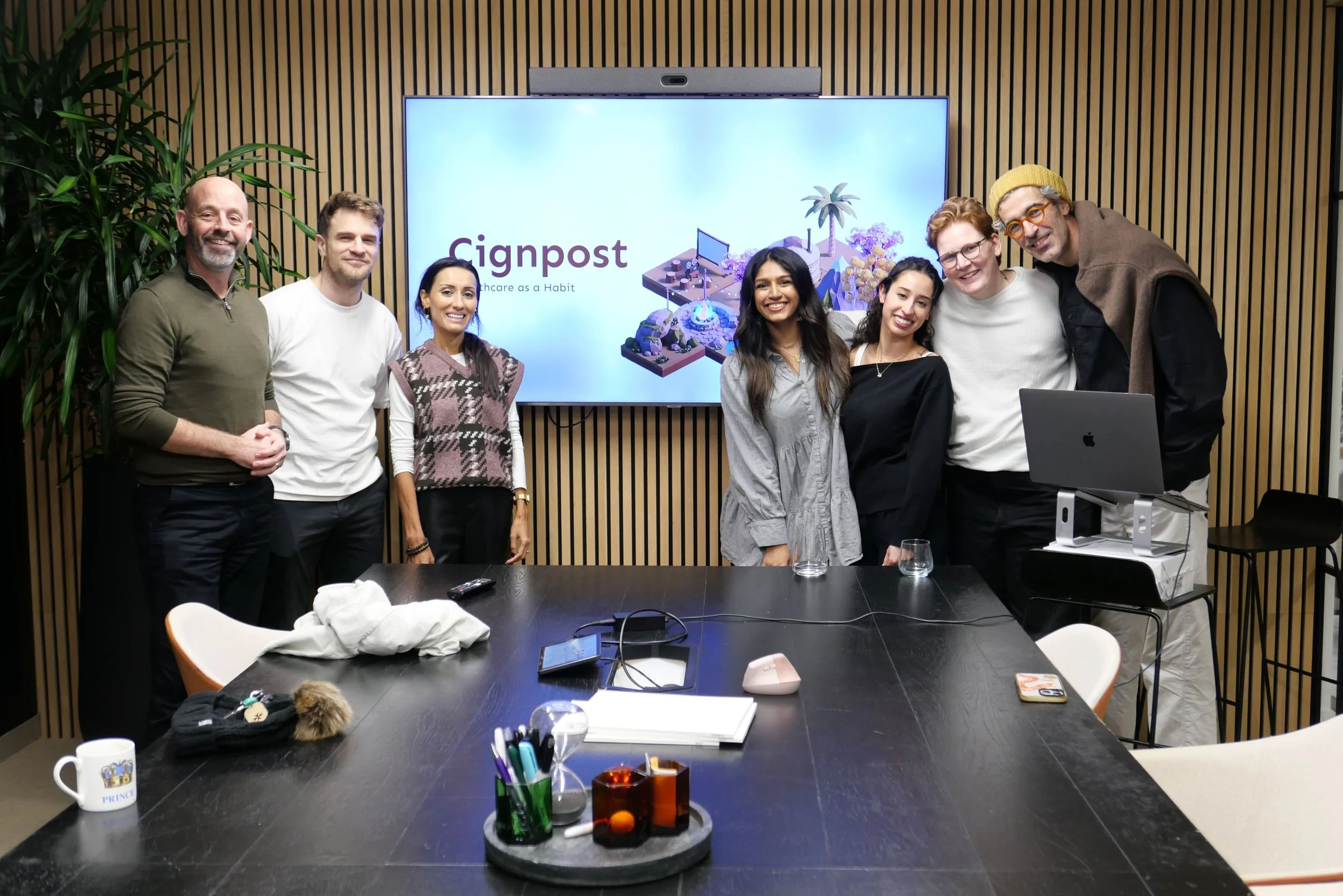 Group of seven people standing in a conference room, smiling, with a large screen behind them displaying the logo 'Cignpost' and text 'Healthcare as a Habit'; a black table in front has various items, including a computer, books, a mug, and glasses.