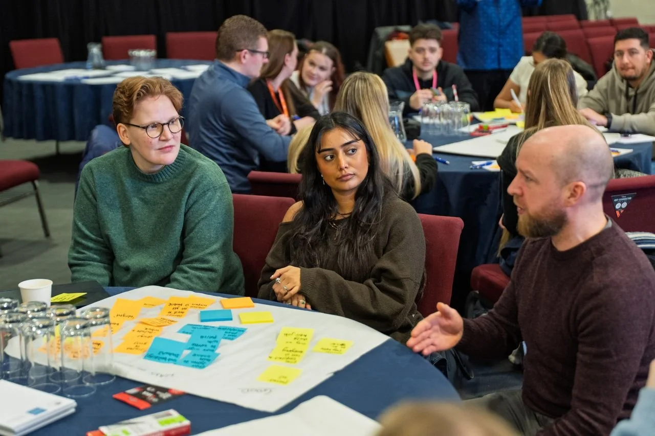 People seated at a round table participating in a workshop or discussion, with colorful sticky notes and papers on the table.