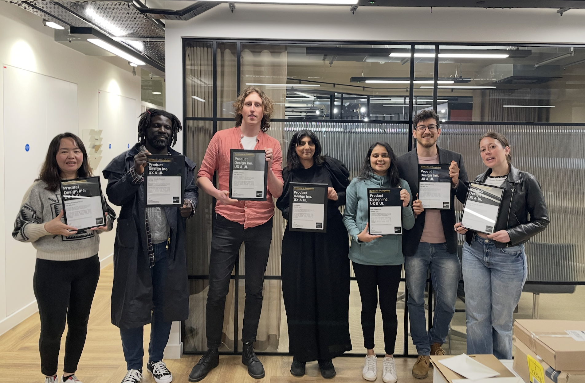 Group of seven diverse people standing in a line inside a modern office, each holding a framed certificate or award.