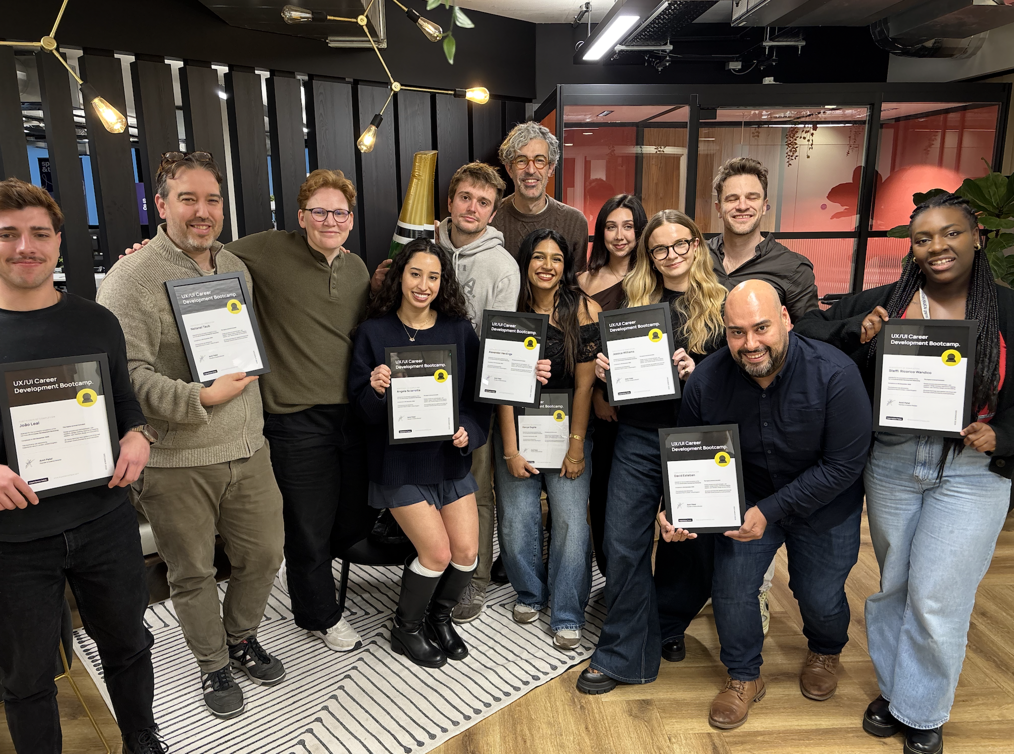 Group of diverse young adults and one older man holding framed certificates at a celebration or graduation event in a modern indoor space.
