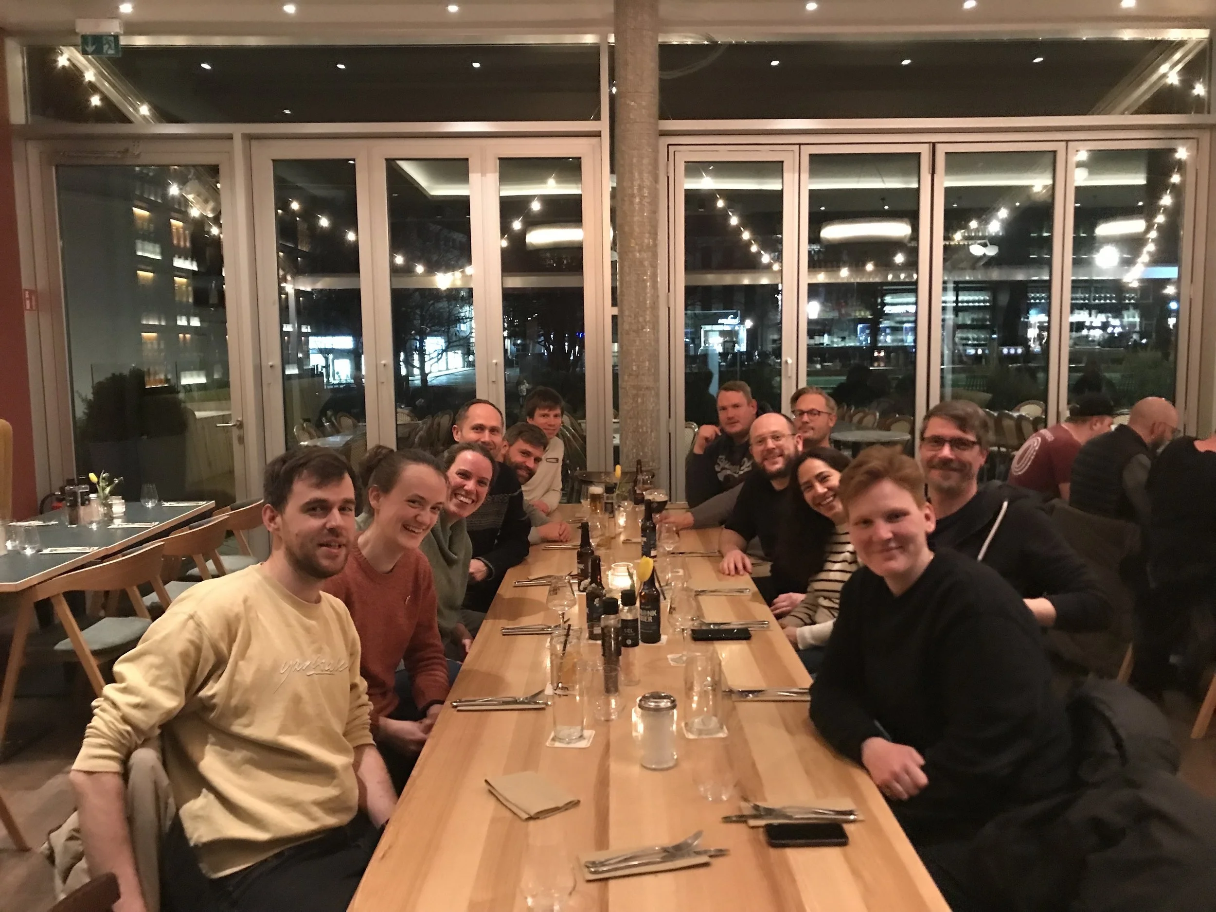 A group of people sitting around a long wooden table in a restaurant, smiling at the camera, with large windows and a cityscape in the background.