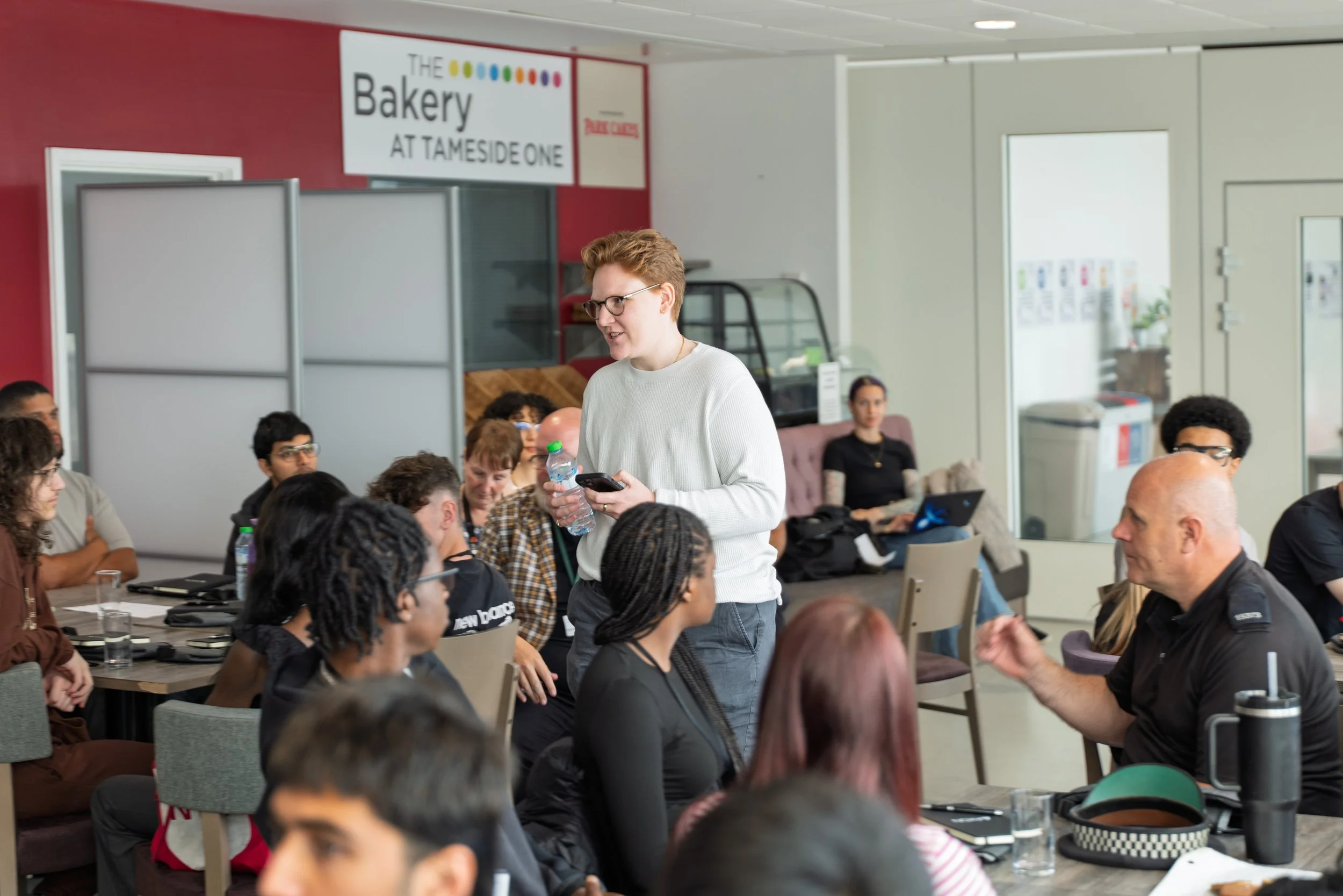 A group of people attending a meeting or workshop in a restaurant or café setting, with a young woman standing and speaking, holding a water bottle and microphone, while others listen attentively.