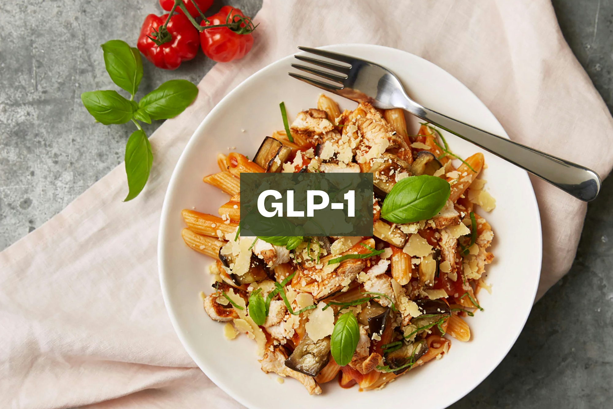 A plate of cooked pasta with vegetables, garnished with fresh basil leaves and grated cheese, with cherry tomatoes and basil on the side. Portion controlled GLP-1 meal.