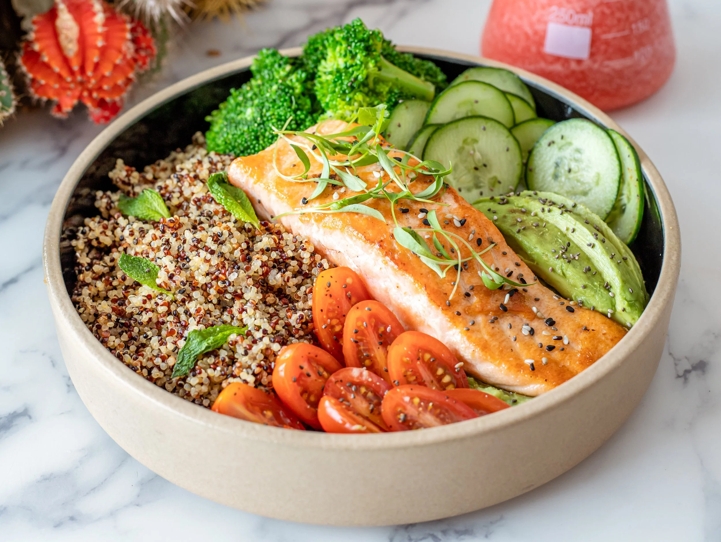 A bowl of food containing cooked quinoa, broccoli, cucumber slices, cherry tomatoes, and a piece of cooked salmon garnished with microgreens and sesame seeds.