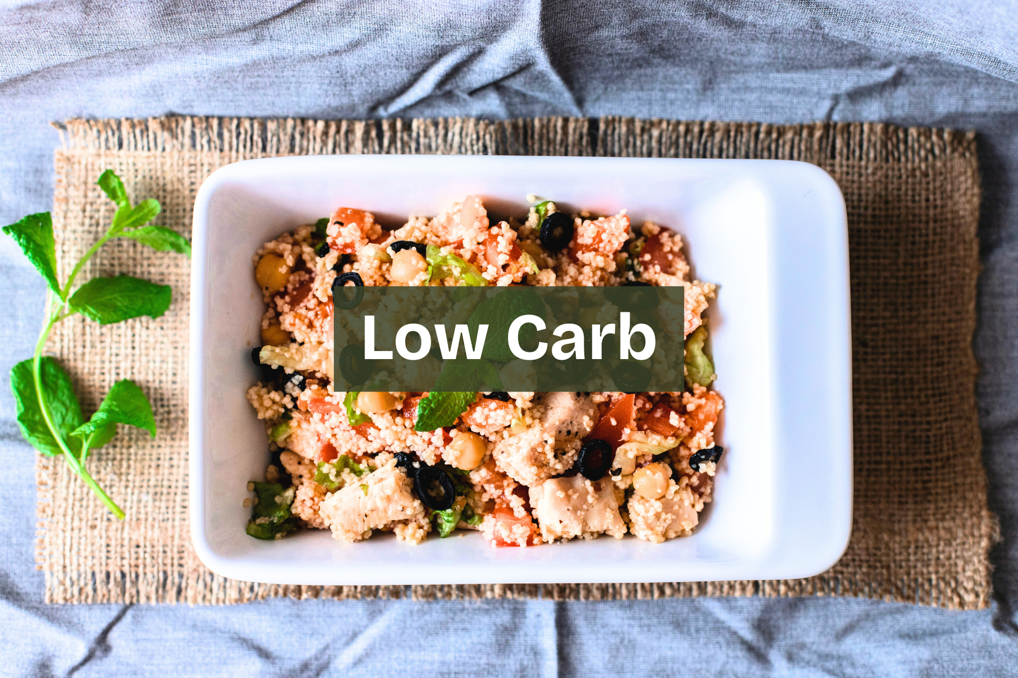A bowl of low carb salad with cauliflower rice, black olives, chickpeas, and chopped vegetables, garnished with a mint leaf, on a woven placemat on a gray table. keto friendly low carb meal with healthy fats Nourix