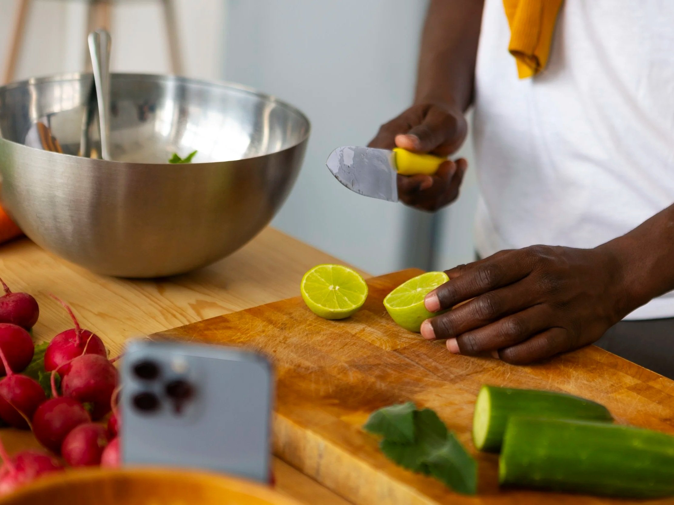 A person slicing a lime with a knife over a wooden cutting board, with radishes and other vegetables nearby, and a metal mixing bowl in the background.