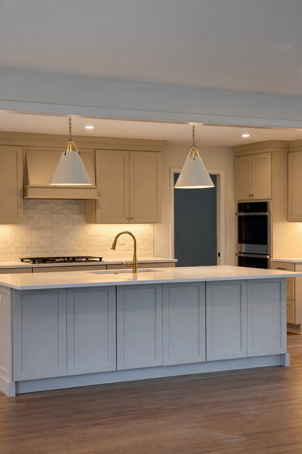 A modern kitchen with a white central island, beige cabinets, brass fixtures, and a white brick backsplash. Pendant lights hang above the island, and the floor is wood.