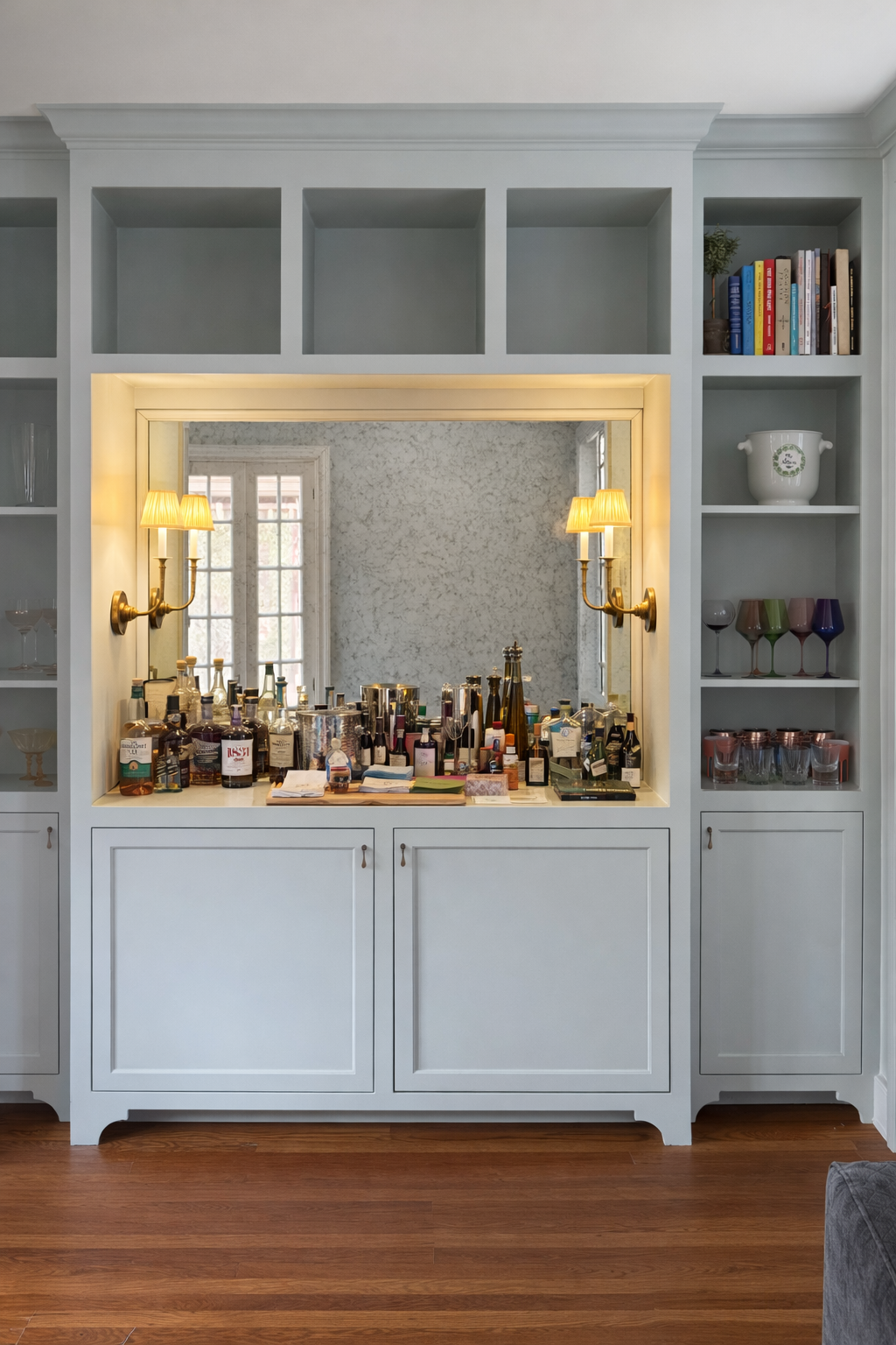 A built-in white cabinet with open shelves on top, containing books and decorative items, and a central mirror with a bar area in front filled with bottles, glasses, and bar accessories, illuminated by wall-mounted lamps.