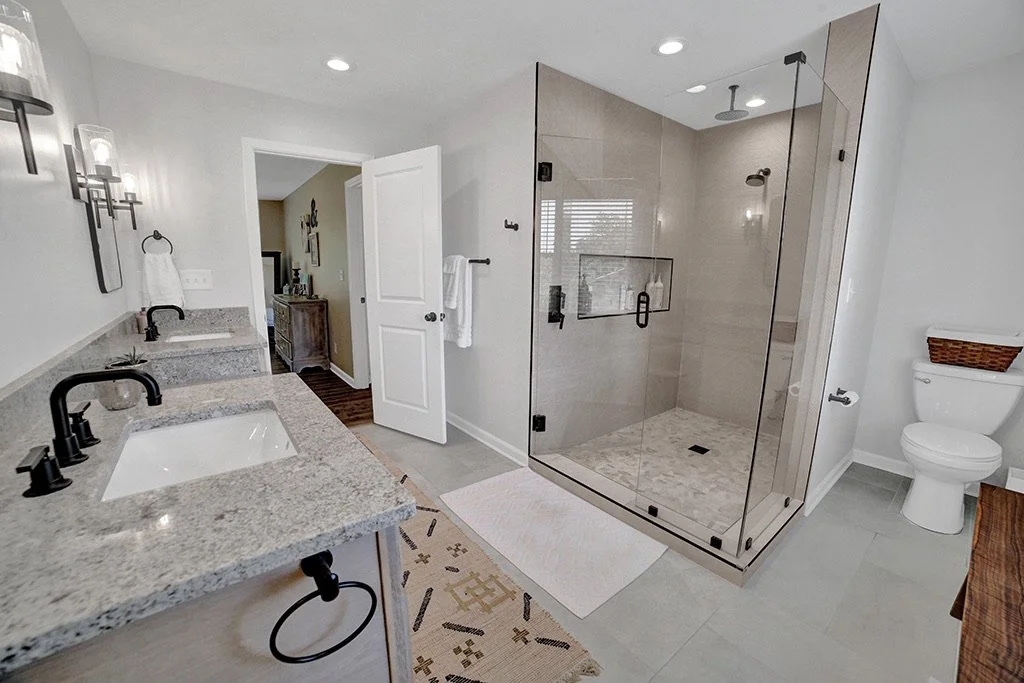 Modern bathroom with dual sinks, granite countertops, black fixtures, a glass-enclosed walk-in shower, a toilet, and a linen basket.
