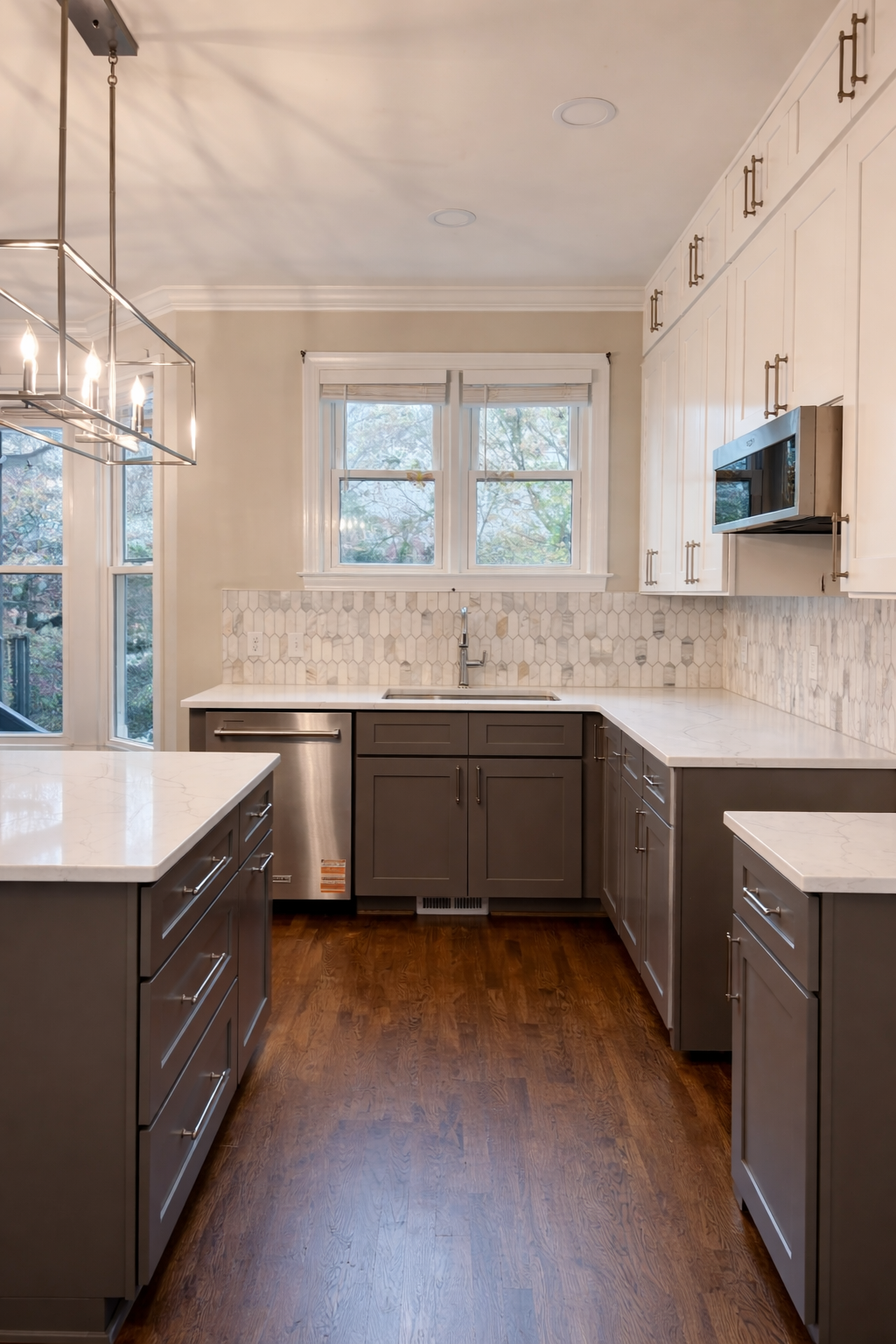 Modern kitchen with white upper cabinets, gray lower cabinets, marble countertops, honeycomb and vertical tile backsplash, wooden flooring, and a window above the sink.