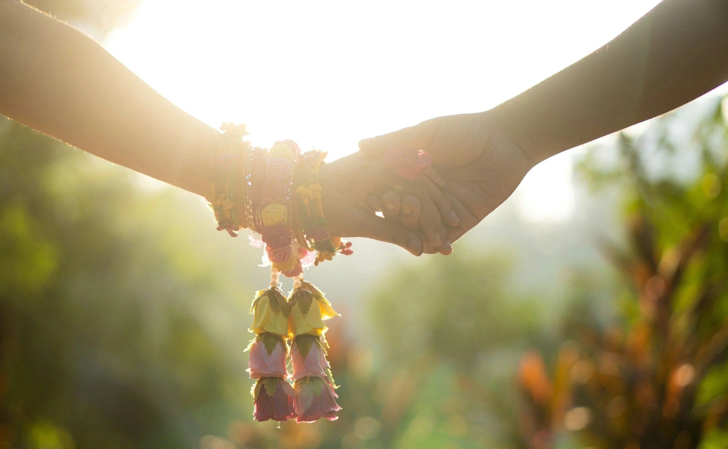 Two hands clasp in a handshake outdoors with bright sunlight shining from behind, and decorative bracelets and flower garlands on the wrists.