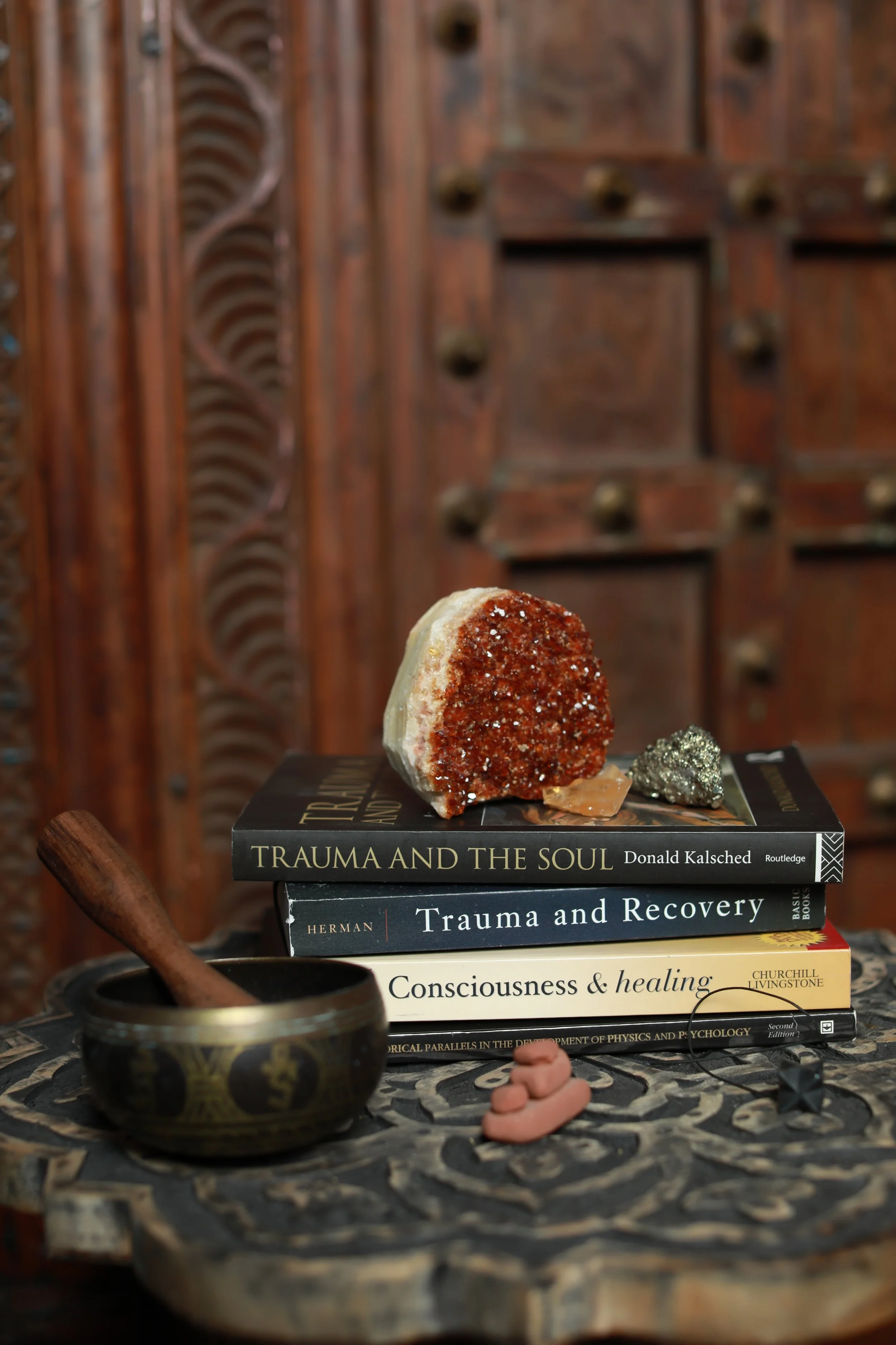 Stack of books about trauma and healing, a medicine bowl with a wooden pestle, a crystal, a mineral, and a small figurine on a decorative table. Resting on top of the books is a geode slice.