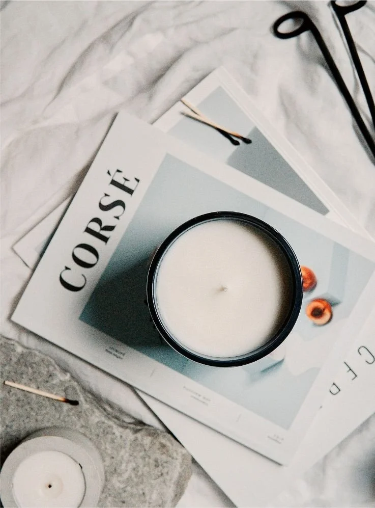 A black candle in a glass jar placed on magazine pages, with a smaller candle on a stone surface, and scattered matches and papers around.