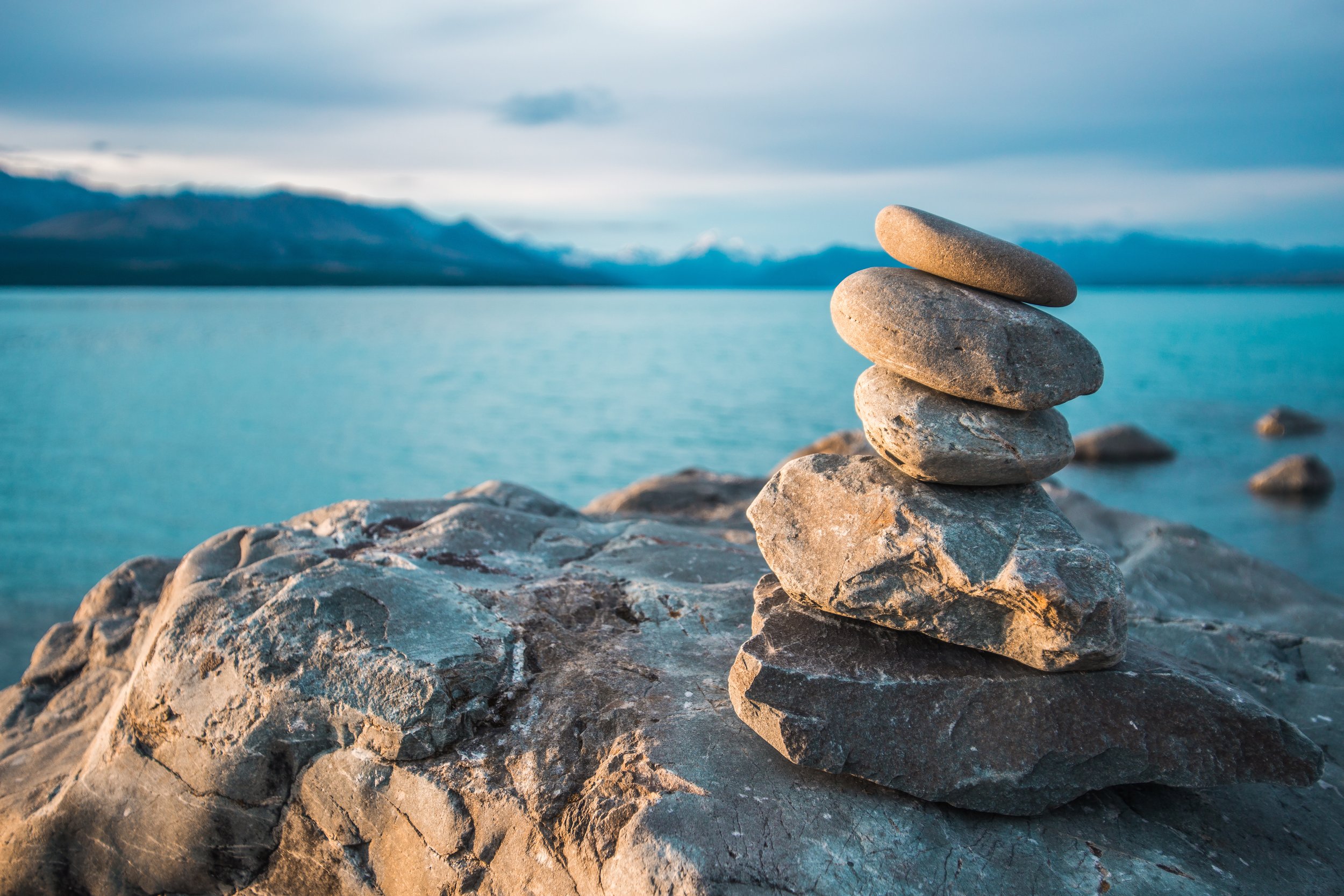 A stack of smooth stones balanced on a rocky surface near a large body of water with mountains in the background under a cloudy sky