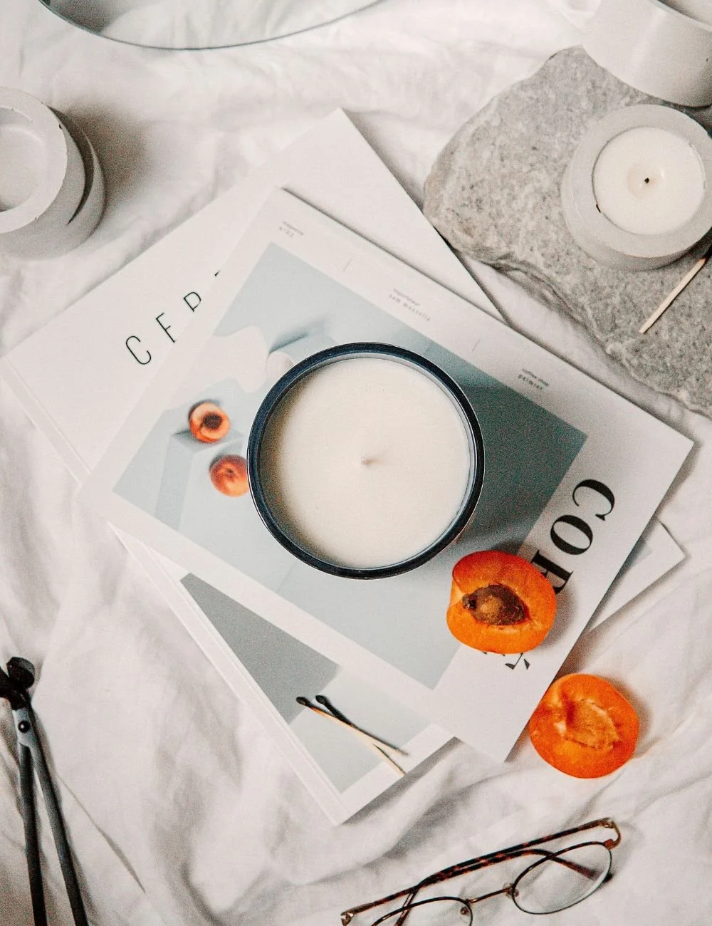 A white candle in a black holder placed on a magazine with a minimalist design, two halved apricots, a small candle on a stone, a pair of glasses, and a clock in the background, all arranged on a white fabric surface.