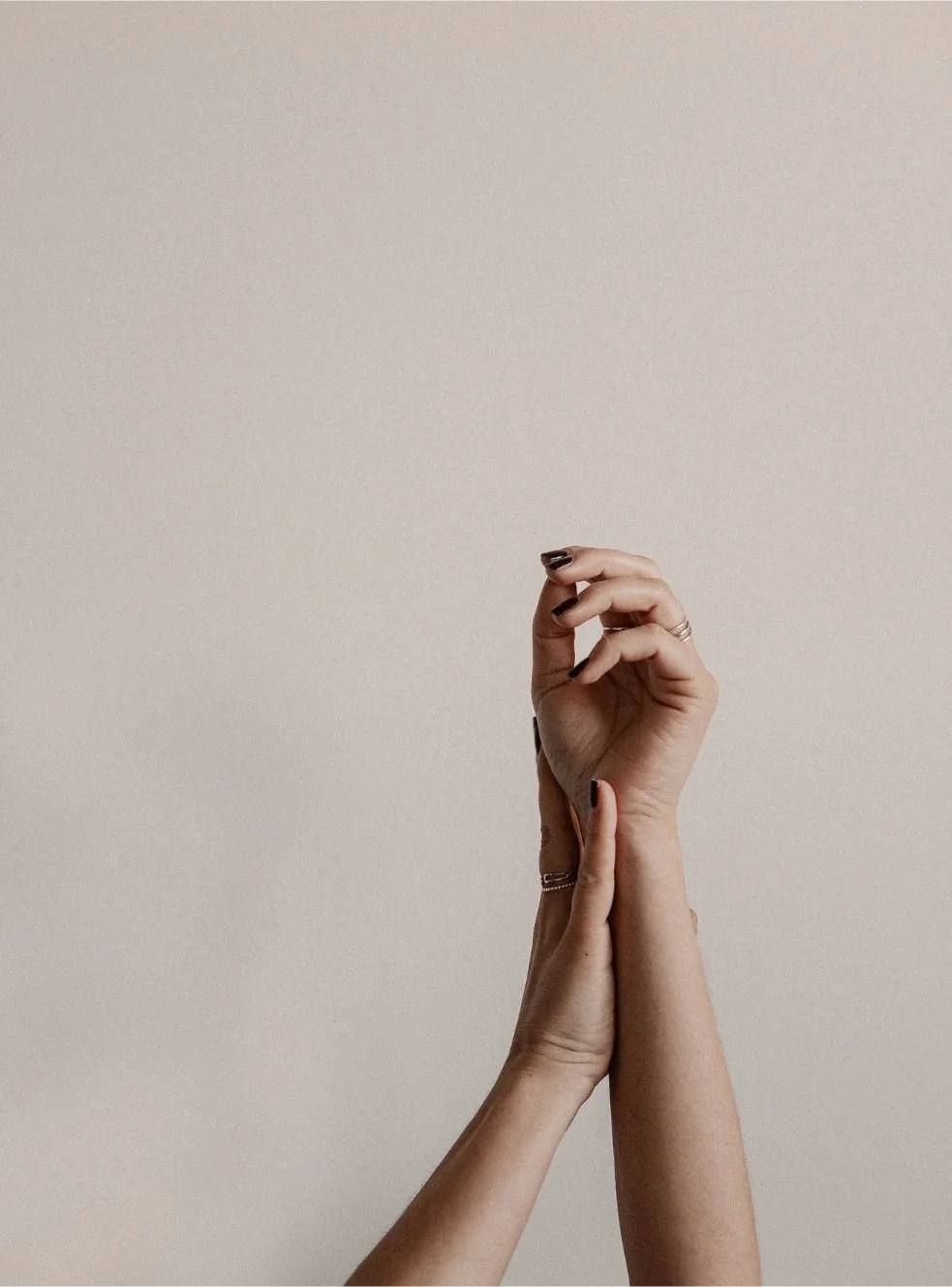 Two hands, one with dark nail polish, gently holding and supporting the wrist of the other hand, against a plain light background.