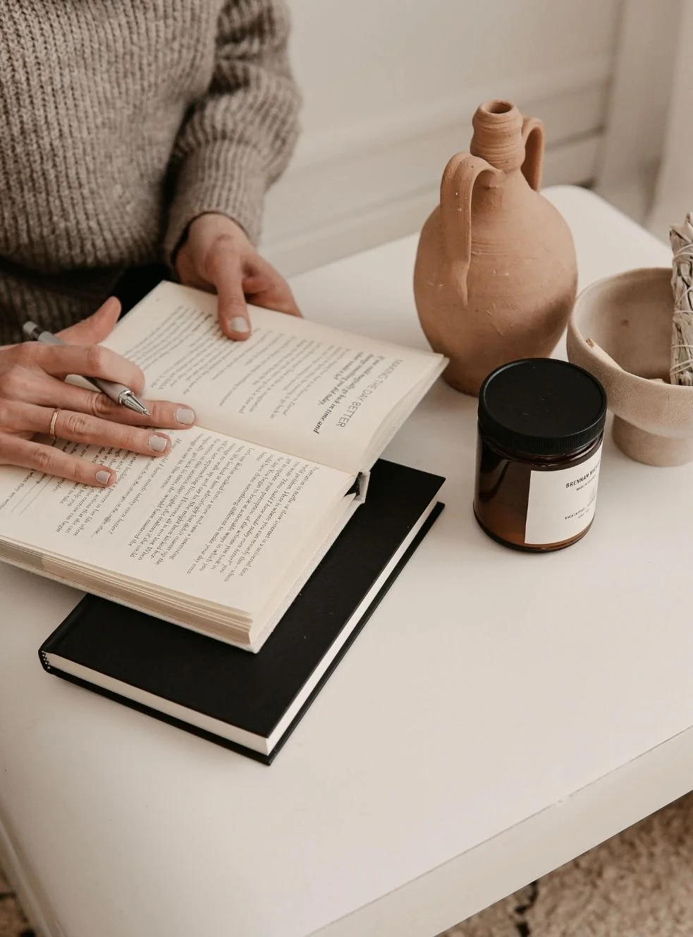 Person in beige sweater reading a book at a white table with pottery vessels, a candle, and a bowl.