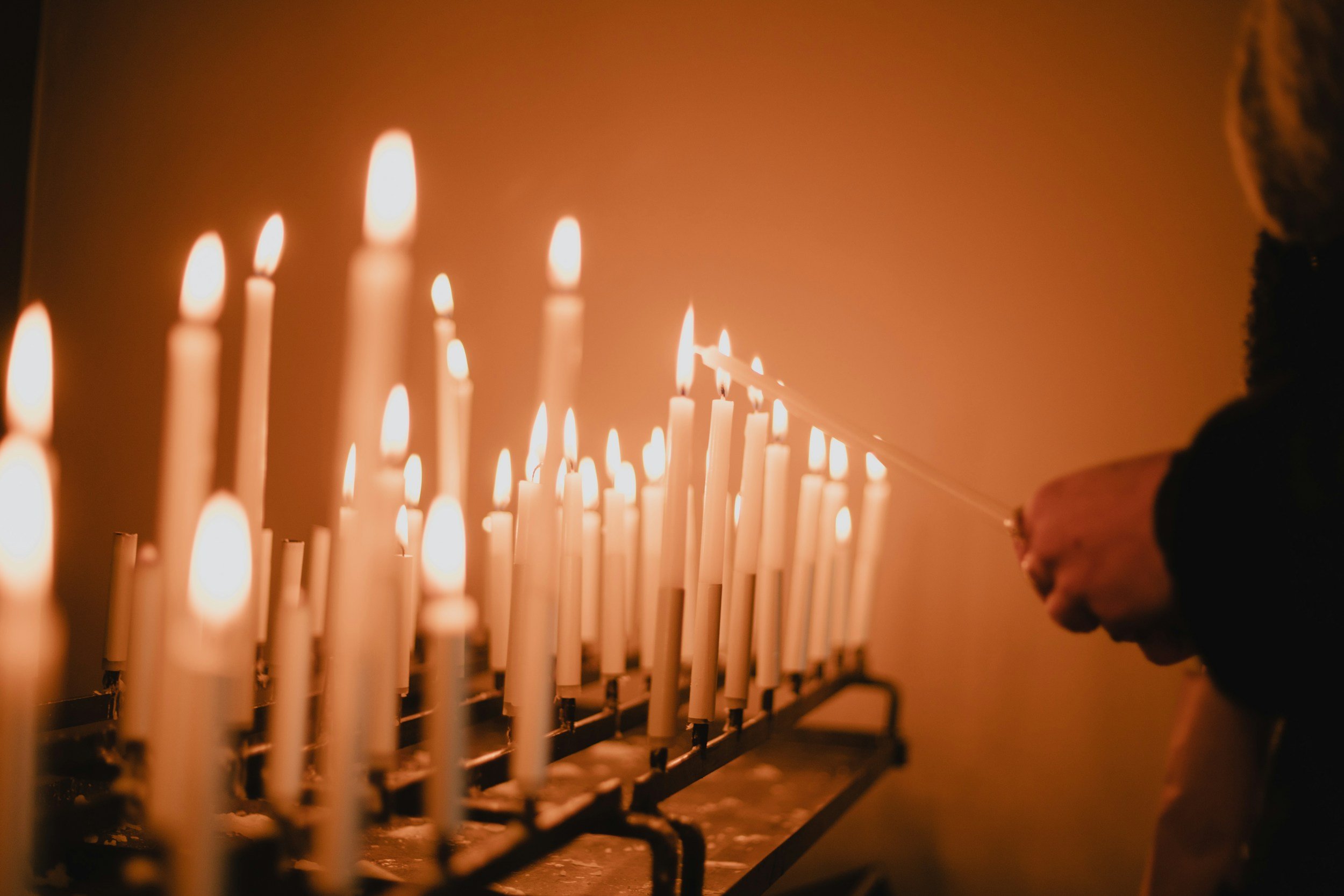 A person lighting a candle from a row of lit candles, with a warm orange glow in the background.