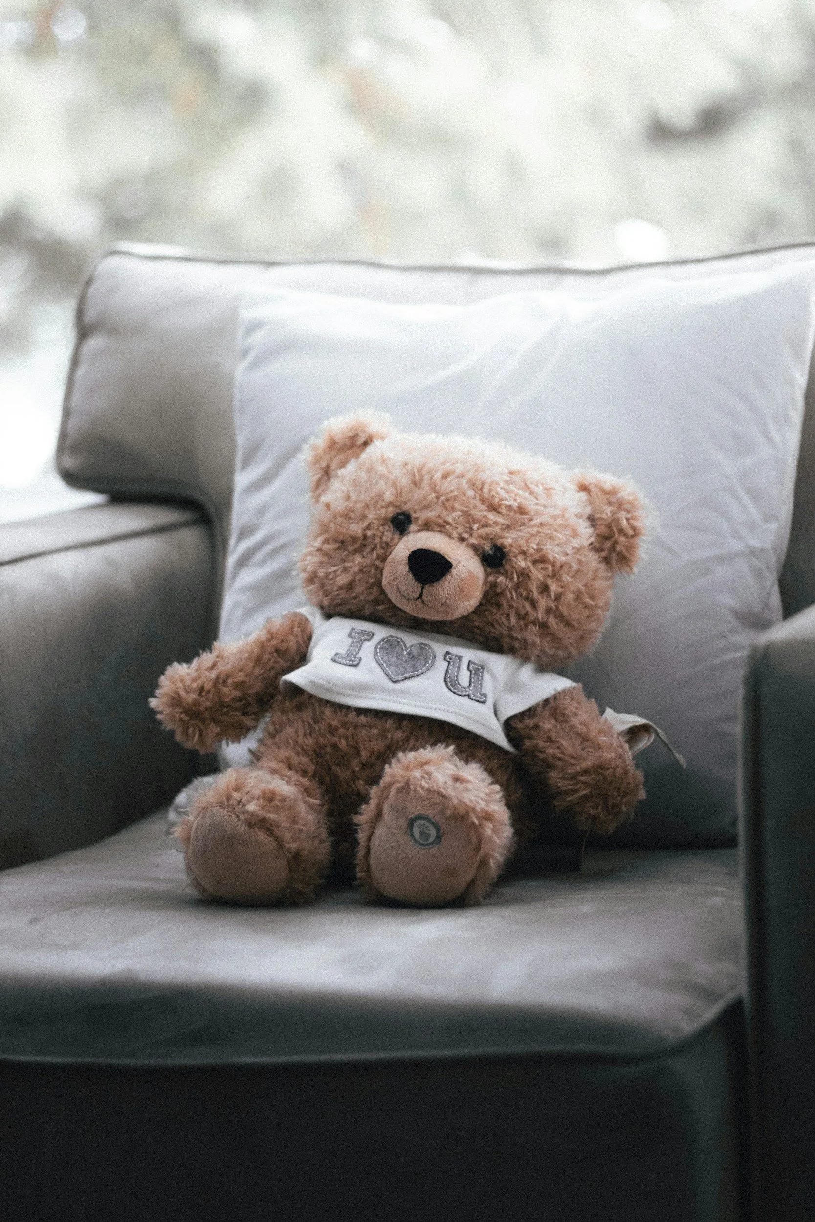 A teddy bear with light brown fur, wearing a white shirt with 'I ♥ U' embroidered on it, sitting on a bed with white sheets and a large white pillow, in a room with soft lighting and neutral colors.