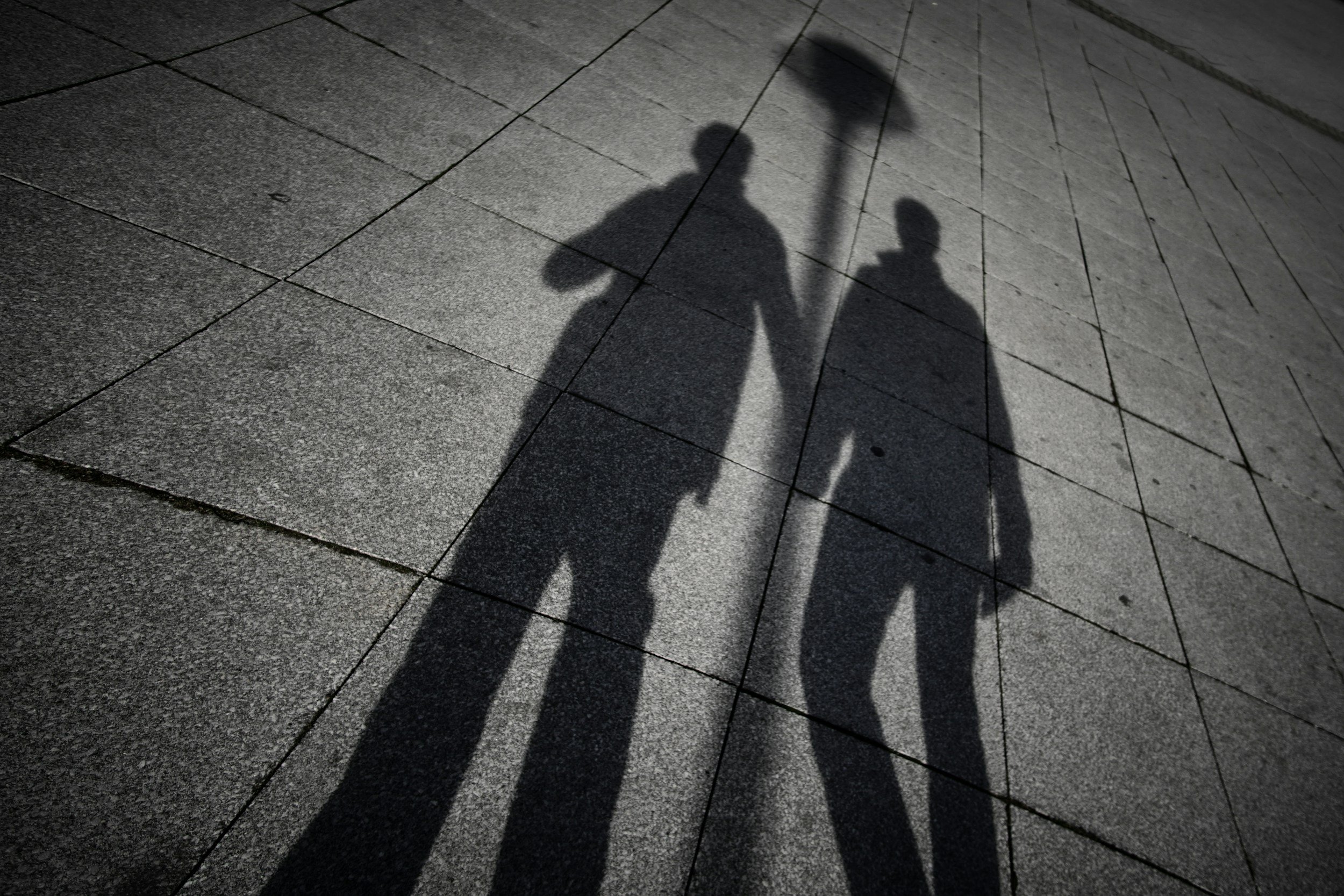 Shadows of two people walking on a tiled sidewalk, with one person carrying a bag and a stick.