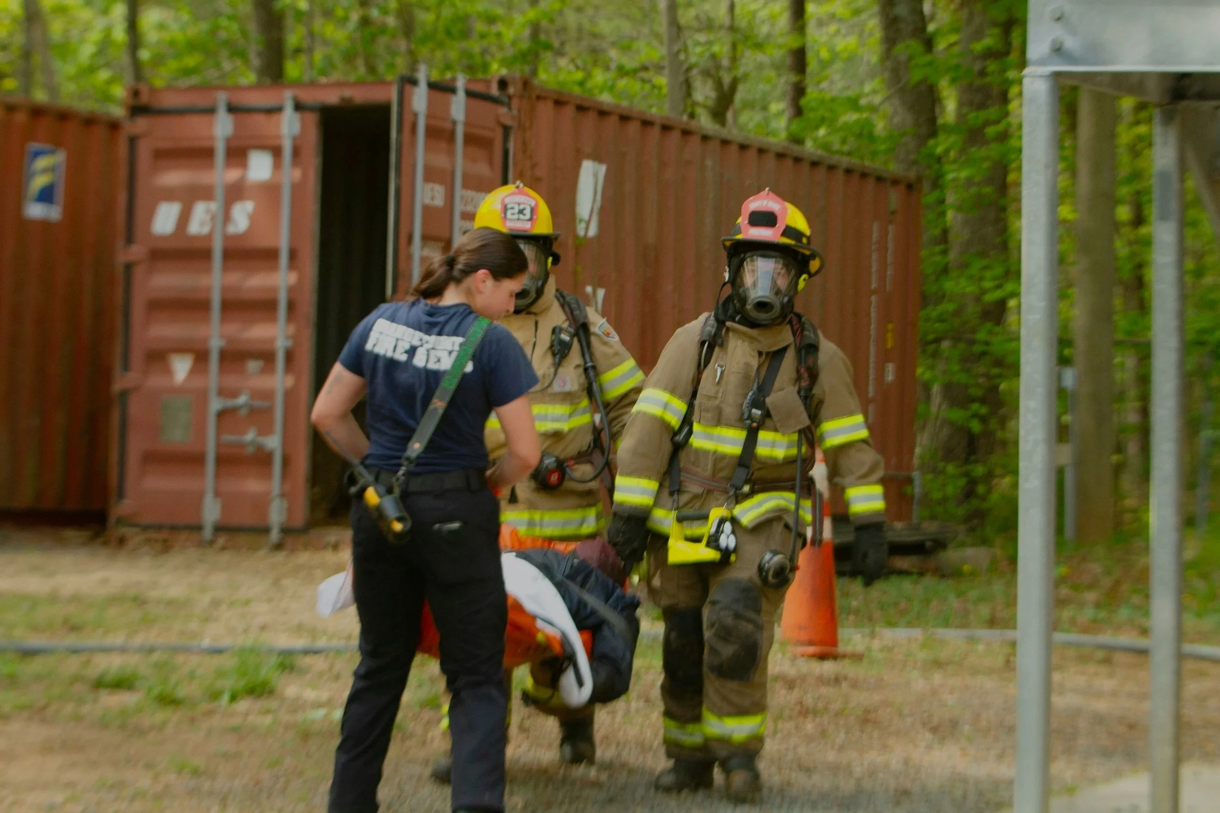 Firefighters assisting a person lying on the ground in a wooded area; a woman with a gear is standing nearby.